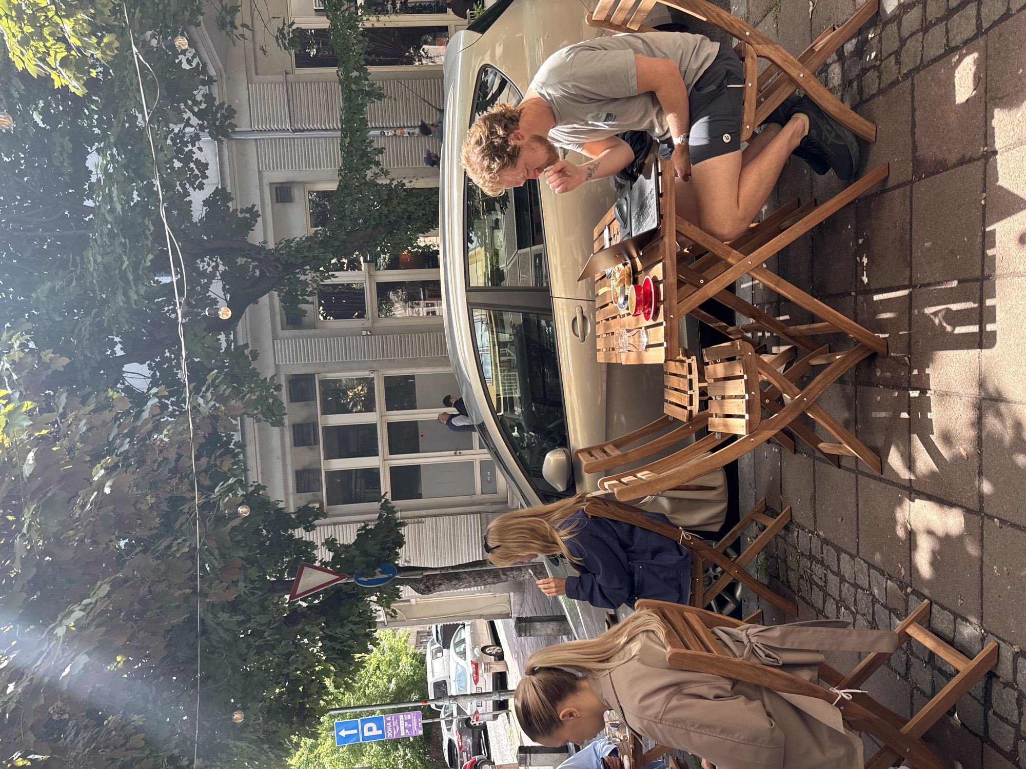 The image shows an outdoor cafe scene in Belgrade, Serbia. Three individuals are seated at wooden tables, with a man leaning in to interact with a laptop and food on the table. A woman in a blue jacket uses a phone, while another woman in a tan coat sits nearby. A light-colored car is parked immediately adjacent to the cafe seating area. The cafe is located on a street lined with buildings, trees, and street signs in the background. Overhead are string lights hanging from the trees.