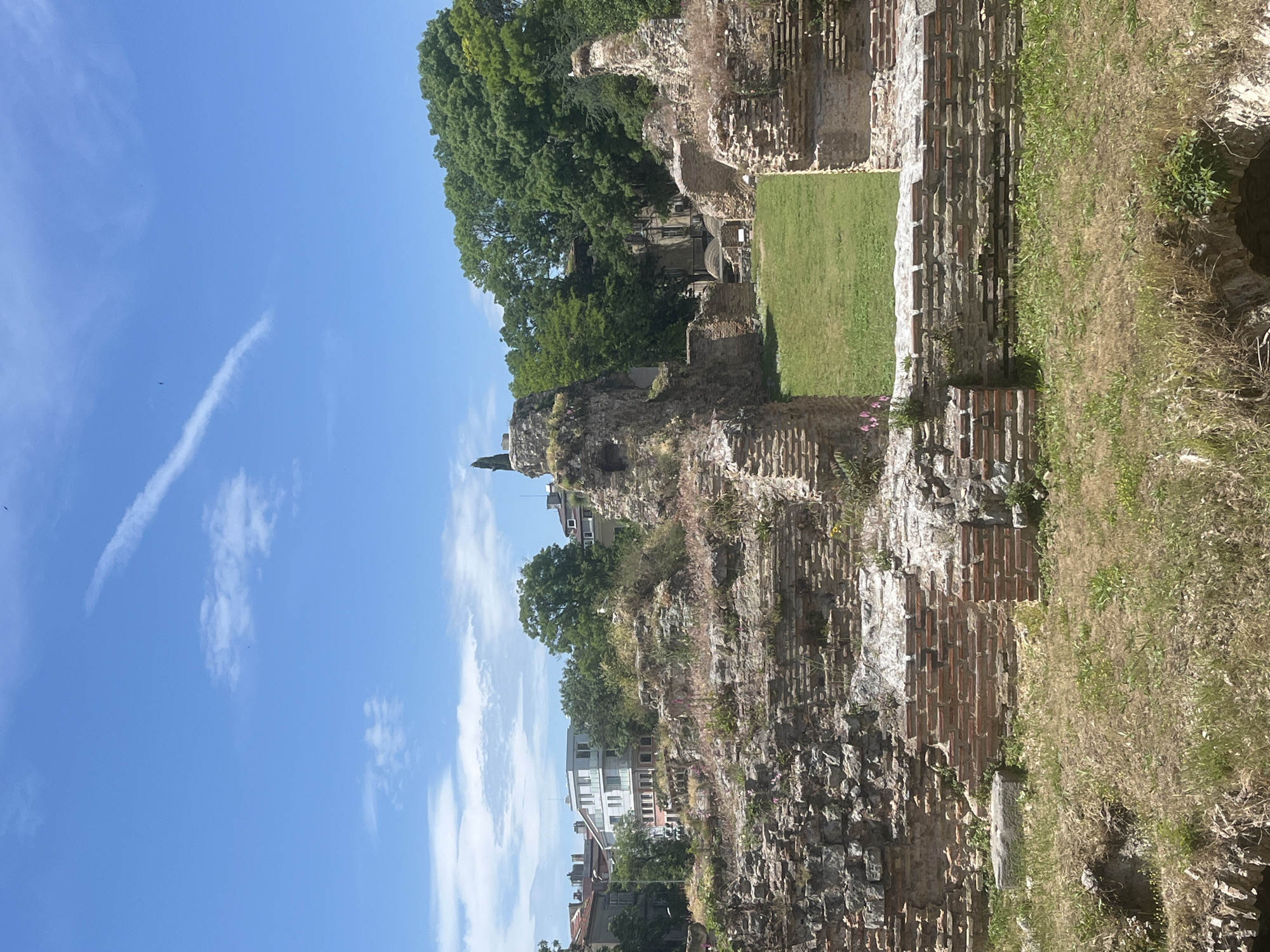 The image captures the remnants of ancient Roman Thermae in Varna, Bulgaria. The partially destroyed brick and stone walls reveal layers of history. A patch of well-maintained green grass contrasts with the weathered stonework. Trees grow behind and within the ruins. In the background, modern buildings provide a juxtaposition of past and present. The sky is clear with a few clouds. No people are visible.