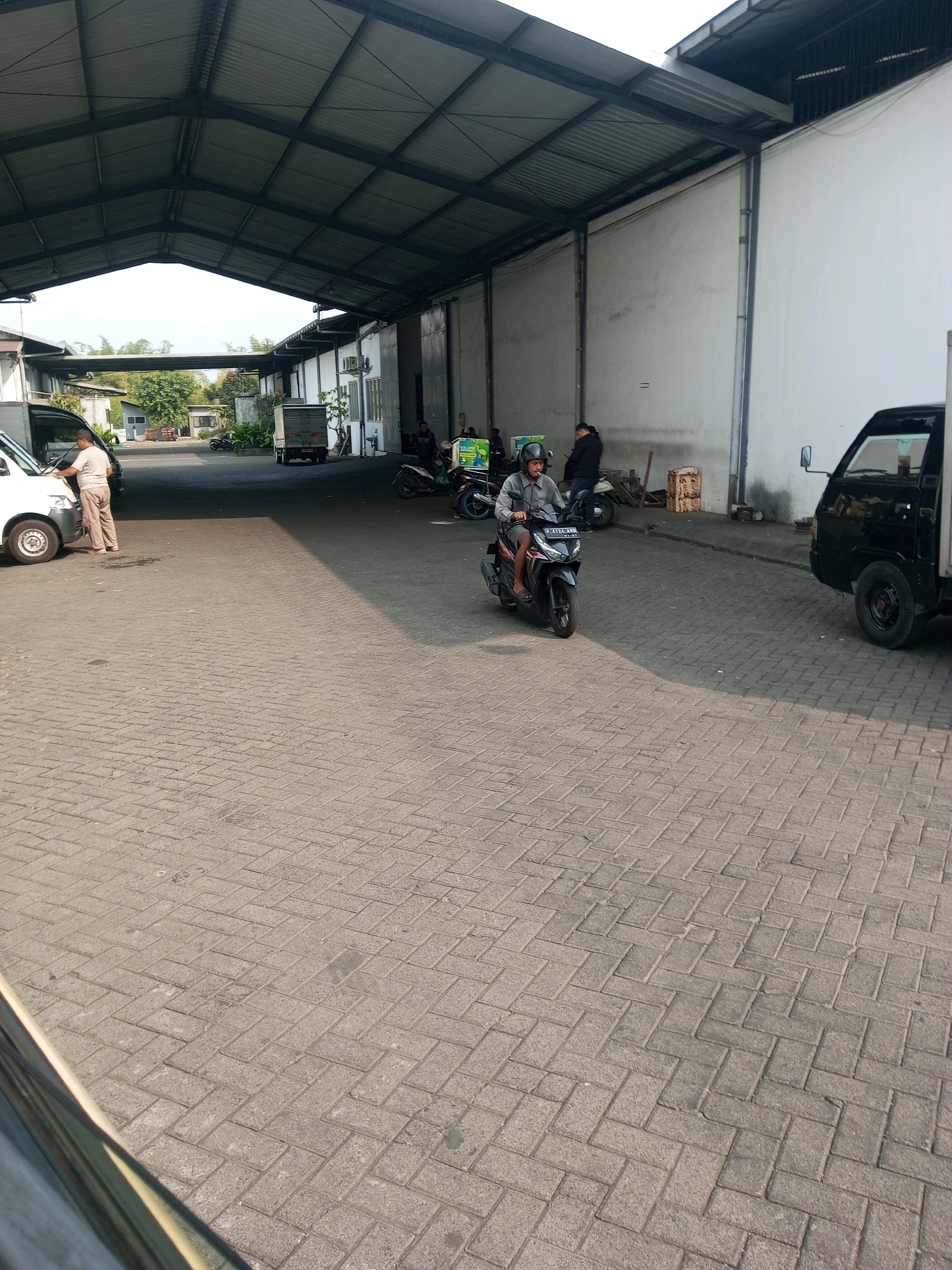 The image shows an outdoor covered area in Mangliawan, Indonesia, likely a warehouse or industrial facility, with a paved cobblestone floor. Sunlight casts long shadows across the ground, indicating it is daytime.
In the foreground, a man wearing a gray shirt and pants, and a black helmet, is riding a black motorcycle. He is looking forward and appears to be smoking a cigarette. The motorcycle has a license plate that reads "B 4174 ABF".
To the left, a white van is parked, and a man in a light-colored shirt and pants is standing next to it, appearing to interact with the vehicle. Further back, several other motorcycles and at least one small truck with a canopy are visible. One of the motorcycles is green and has a box attached to the back, suggesting it might be used for delivery. Two individuals are standing near this motorcycle.
The background features the corrugated metal roof of the covered area, supported by metal beams and poles. The walls of the adjacent buildings are white. Trees and other vegetation are visible beyond the structures. The overall environment appears to be a functional, possibly busy, commercial or logistical space.