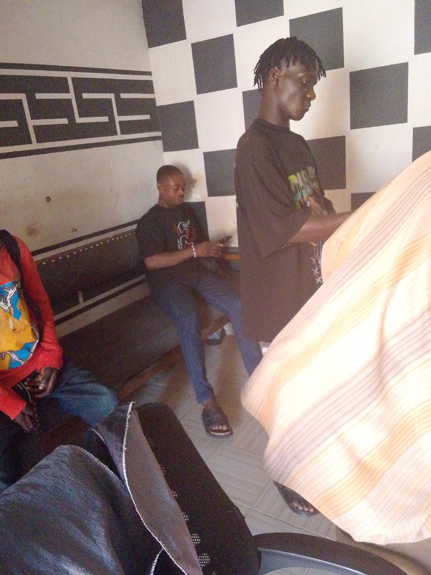 This image depicts an indoor scene featuring three men in what appears to be a waiting area or communal space in Jos, Nigeria.
On the right, a man with dark skin and dreadlocks stands facing partially right, looking downwards. He wears a black t-shirt with colorful, indecipherable graphics and black sandals. His hands are clasped in front of him, and a large, light-colored fabric with subtle vertical stripes, possibly a curtain or draped garment, obscures the lower right portion of the frame, extending towards him.
In the center, another man with dark skin and short hair is seated on a dark, possibly upholstered, bench. He wears a black t-shirt with a white, unreadable design, blue jeans, and dark sandals. He is engrossed in a mobile phone he holds in his hands.
On the far left, a third man is partially visible, also appearing to be seated. He wears a red long-sleeved shirt featuring a prominent yellow and blue graphic design, possibly depicting an antler-like or tree-like motif. His lower body is mostly obscured by a dark bag or object in the immediate foreground.
The room's walls are distinctly decorated. The wall behind the seated man has a light background with a bold, black geometric border pattern, reminiscent of a Greek key design. The corner wall to the right features a contrasting black and white checkerboard pattern. The floor is tiled with light-colored squares. The scene suggests a calm, casual atmosphere, likely during the daytime. No legible text is visible on clothing or in the environment.