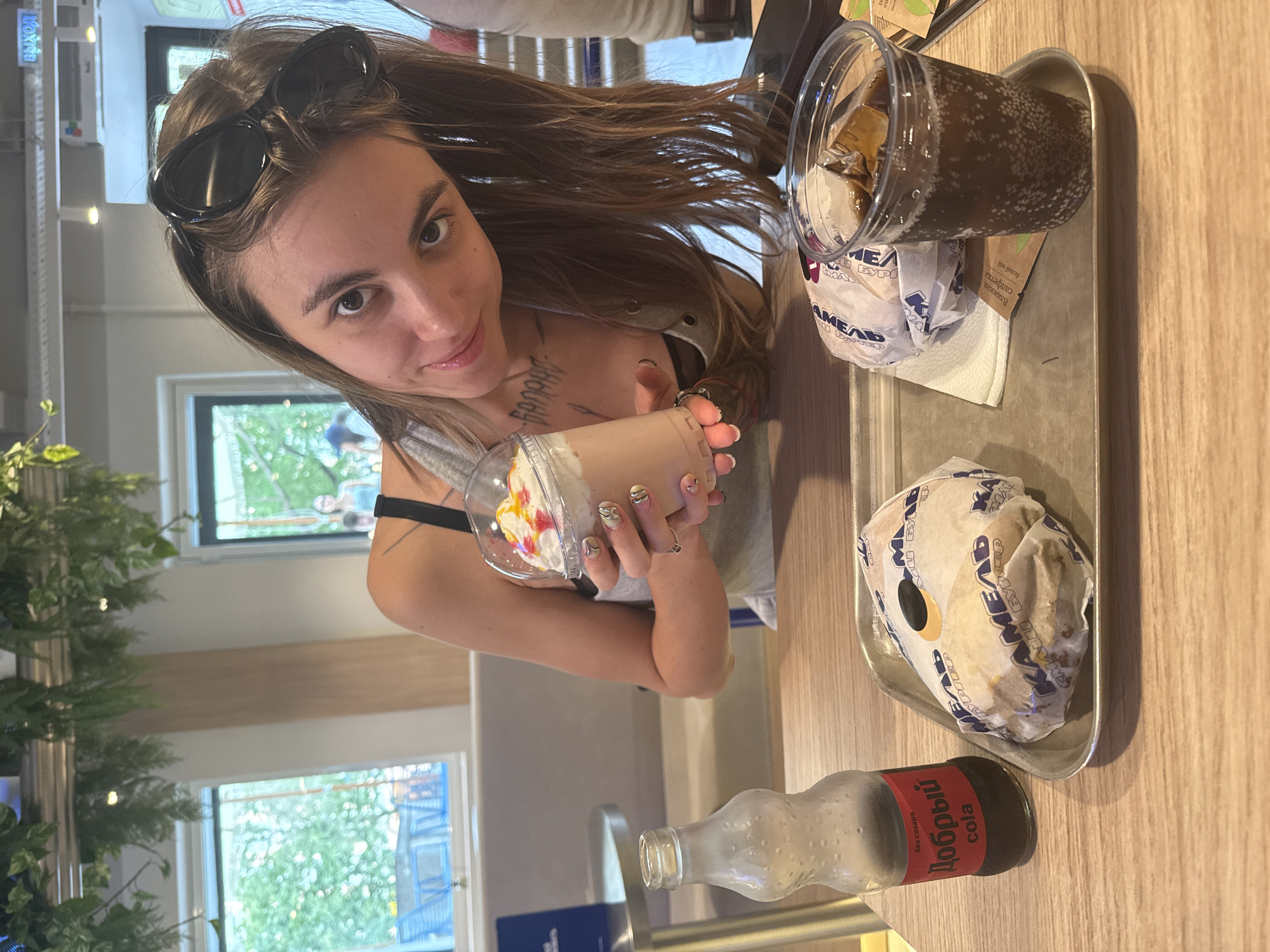 A young woman with long brown hair and sunglasses perched on her head leans forward into the frame, holding a cup with whipped cream and colorful toppings. She is wearing a grey tank top with a black bra underneath, and has tattoos on her chest and arm. In front of her on a metal tray are two burgers wrapped in paper, a glass bottle with a red label that says "Добрый cola", and a clear plastic cup filled with ice and a dark brown beverage, likely cola. The scene appears to be indoors, possibly a fast-food restaurant, with large windows in the background showing greenery outside. A sign above a doorway in the background reads "ВЫХОД" (Exit in Russian). The setting suggests a casual dining environment in Moscow, Russia.