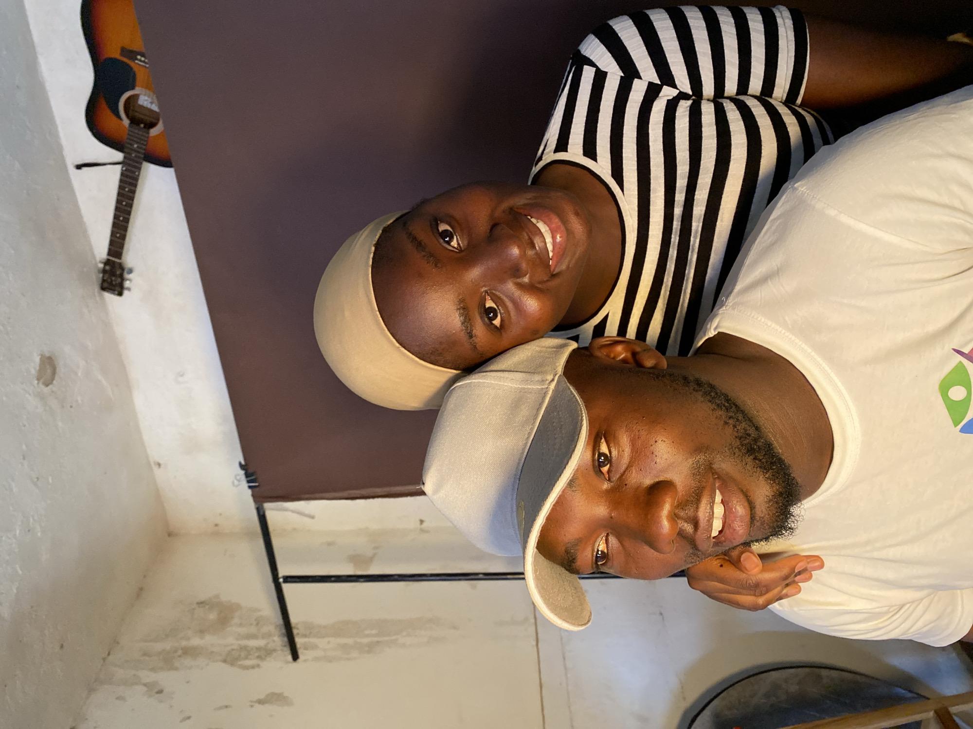 An indoor portrait displays two individuals, likely a man and a woman, posing together with happy expressions. The image is rotated 90 degrees clockwise.
The woman is positioned above the man (when the image is viewed upright), wearing a light-colored head covering and a black and white striped sleeveless top. She has dark skin and is smiling broadly towards the camera, her eyes bright.
The man is positioned below the woman, wearing a light-colored baseball cap and a white short-sleeved t-shirt. He has a dark beard and is also smiling, looking towards the camera. A colorful, abstract logo is visible on the right shoulder area of his white t-shirt, but no text is discernible.
The setting appears to be a casual indoor space. In the background, on the far left, a classical acoustic guitar with a brown body and dark neck hangs against a white or off-white wall that shows some signs of age or texture. A large, dark brown or purplish panel serves as a backdrop behind the individuals. The floor or lower wall area, visible beneath the man, is light-colored and also shows some marks. A dark, circular object is partially visible in the bottom right corner. Both individuals convey feelings of warmth and happiness through their smiles. The location context is Vom, Nigeria.