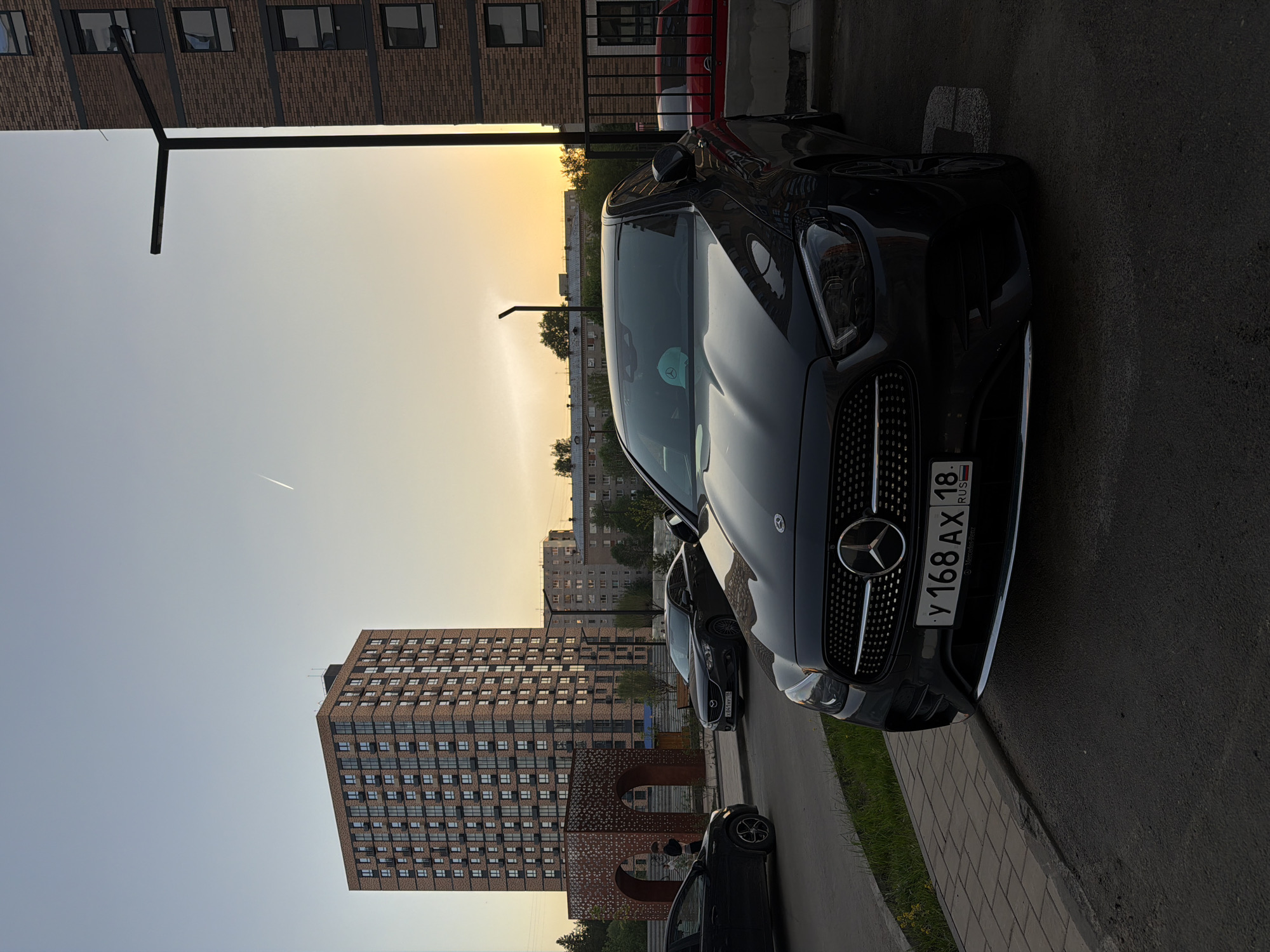 The image shows a black Mercedes-Benz parked on a paved surface next to a curb with grass, with a view of a residential area at sunset. The car's license plate, "У 168 АХ 18 RUS," indicates it is registered in the Udmurt Republic of Russia. In the background, there is a tall, multi-story brick building with many windows, along with other residential buildings of varying heights. The sky exhibits colors associated with sunset. The location is possibly in the city of Izhevsk, Russia, given the license plate registration of the car. There are also other cars parked nearby, though they are less prominently featured.