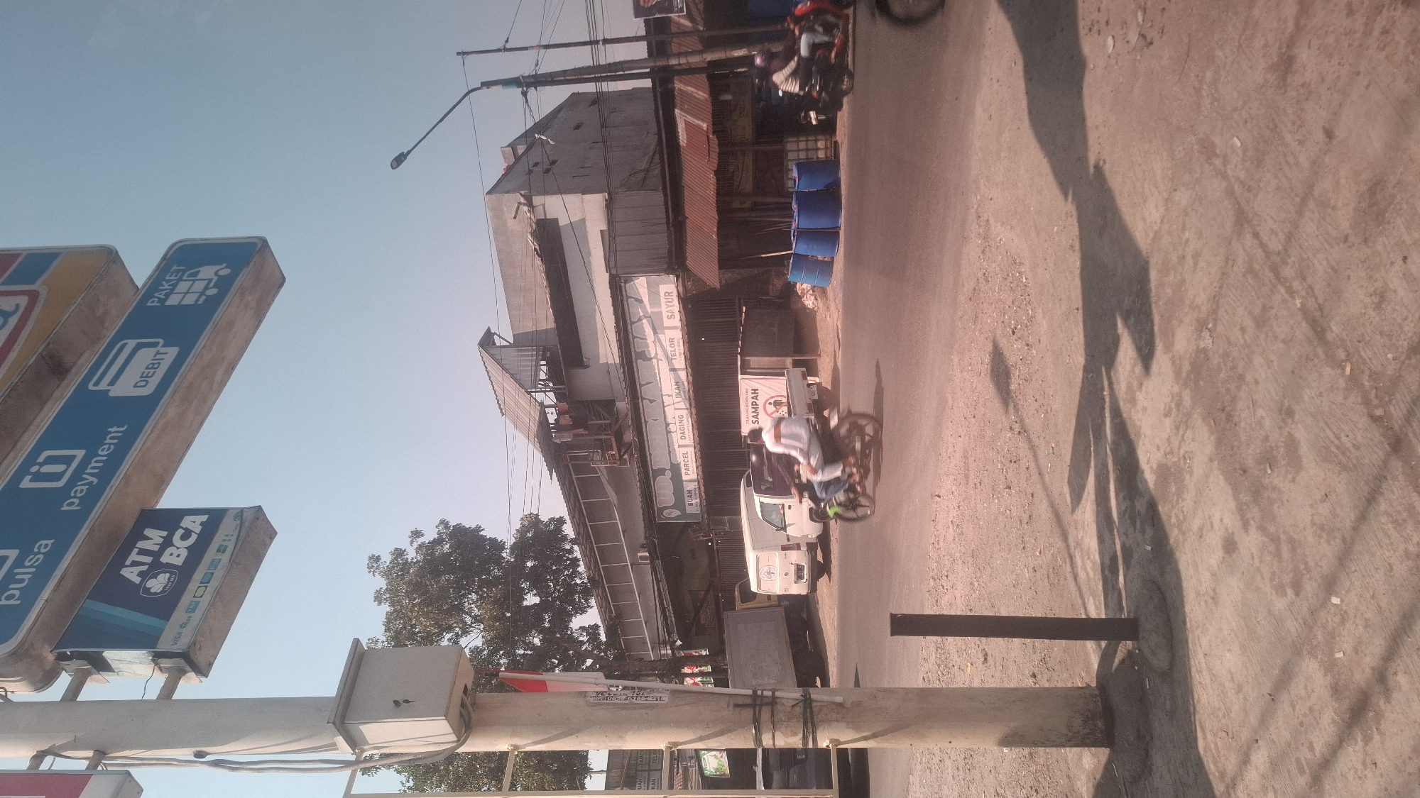The image captures a street scene in Rancagoong, Indonesia, characterized by a clear sky and bright sunlight casting distinct shadows. In the foreground, a light-colored utility pole stands on the left, with a small grey electrical box attached to it. A dark, slender pole is planted next to it in the dirt. The road surface is a mix of asphalt and gravel.
Across the street, a multi-story building complex is visible. A large blue and white sign with text and icons for "pulsa," "payment," "ATM BCA," "DEBIT," and "PAKET" is prominently displayed on the left side of the building's facade. Below this sign, another banner advertises various items: "BUAH," "PARCEL," "DAGING," "IKAN," "TELOR," and "SAYUR."
Several vehicles are present. A white delivery truck is parked on the left. To its right, a smaller white truck is stopped, displaying a "SAMPAH" (trash) sign. In front of the smaller truck, a green motorcycle carrying two individuals is in motion, heading towards the right side of the frame. Further to the right, another motorcycle with a rider is also moving. A row of blue barrels is stacked on the ground in front of a section of the building with a rusty corrugated metal roof. The background shows trees and the sky.