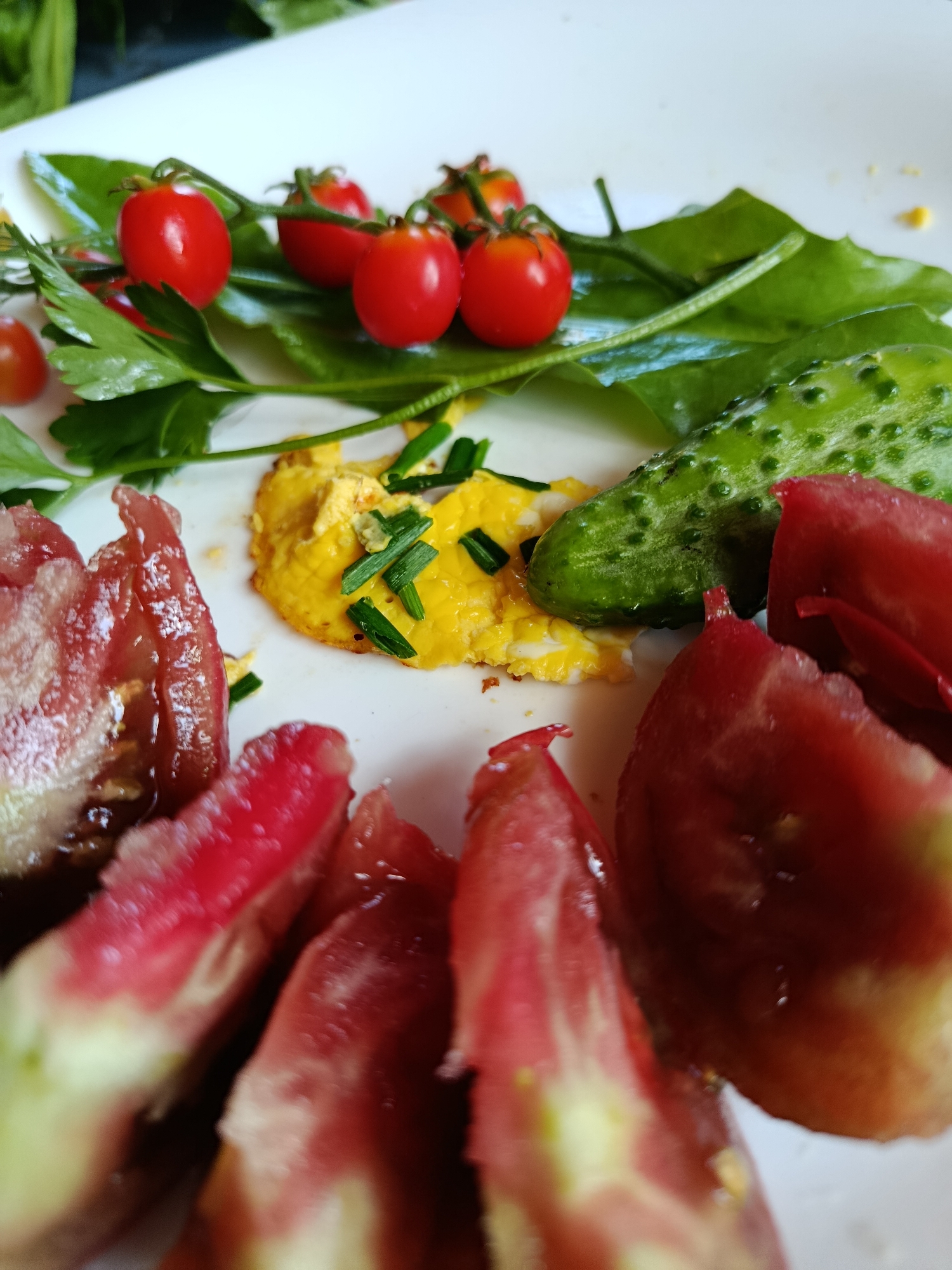 A close-up shot displays a white plate with a meal. The plate holds a small pile of scrambled eggs topped with chopped chives, a sliced cucumber, cherry tomatoes on the vine, and large, roughly chopped pieces of heirloom tomatoes. The background is blurred, showing hints of green leaves and a dark surface. There are no people or discernible urban elements present to identify a specific location in Verea, Russia.