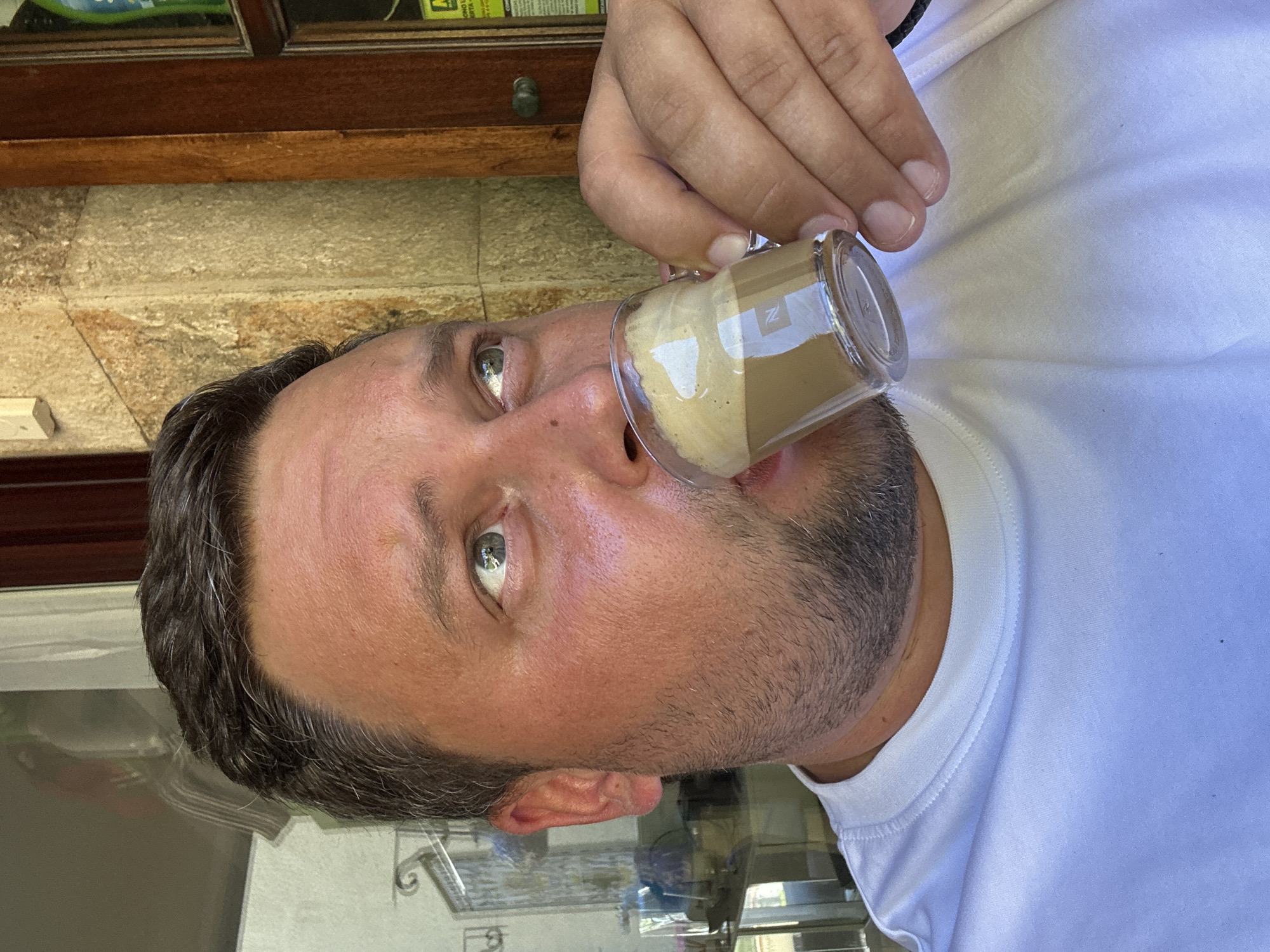 A man is drinking from a small, clear glass cup that contains a beverage that appears to be a type of coffee drink, potentially a latte macchiato, based on the layering of a lighter frothy liquid and a darker coffee layer. The glass has a "Z" design. He is wearing a white t-shirt. The background includes a stone wall, a wooden window frame, and a blurry view of what appears to be an outdoor setting. Based on the location, Sant Feliu de Guíxols, Spain, this scene is likely taken at an outdoor cafe or restaurant.