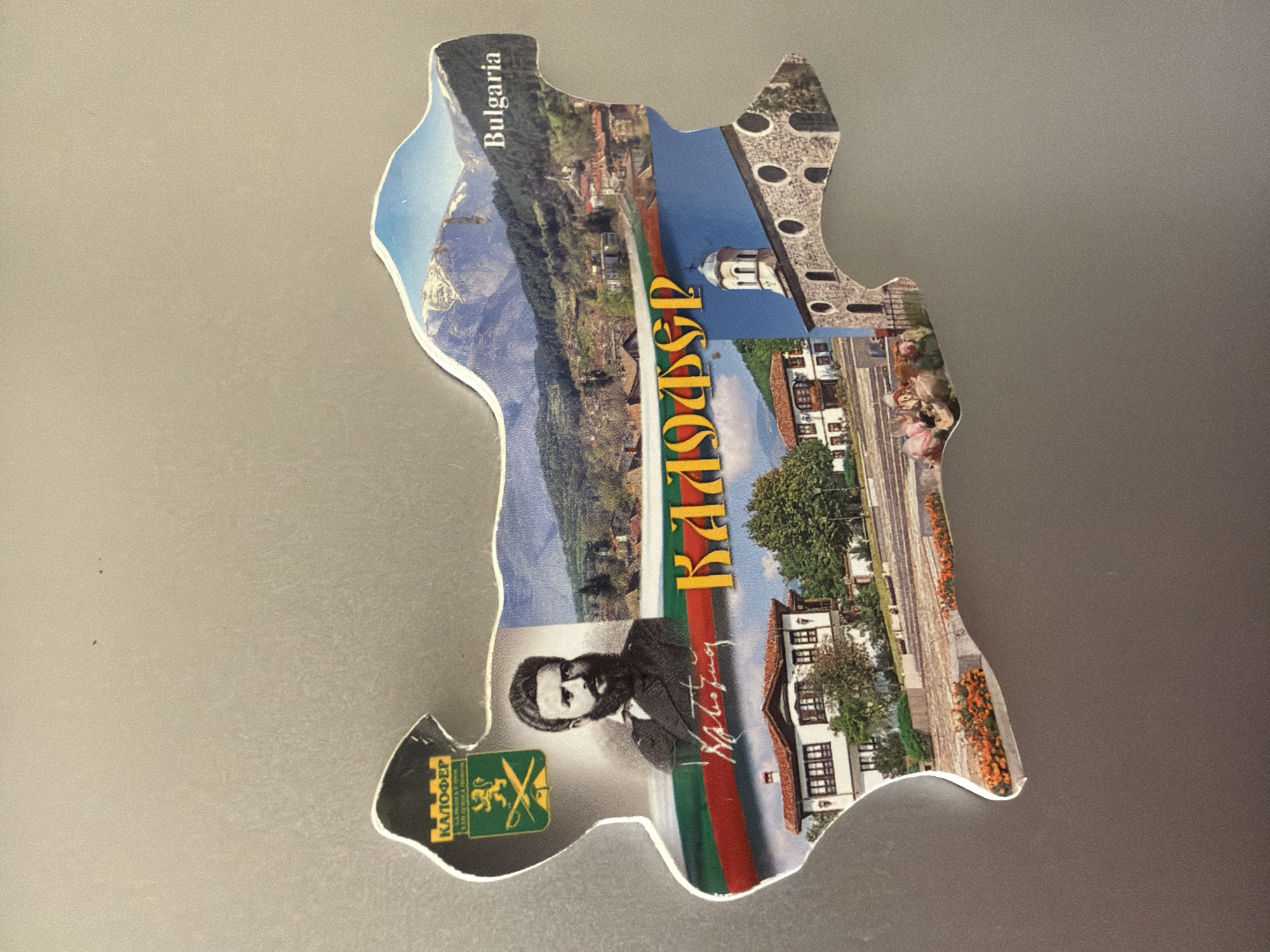 The image is of a magnet shaped like Bulgaria. The magnet features several images: a portrait of a man with a beard, a crest with a lion and swords, a cityscape with mountains in the background, a building with a red roof, a church tower, and the text "BULGARIA" and "КАЛОФЕР". The magnet is placed on a metallic surface.
