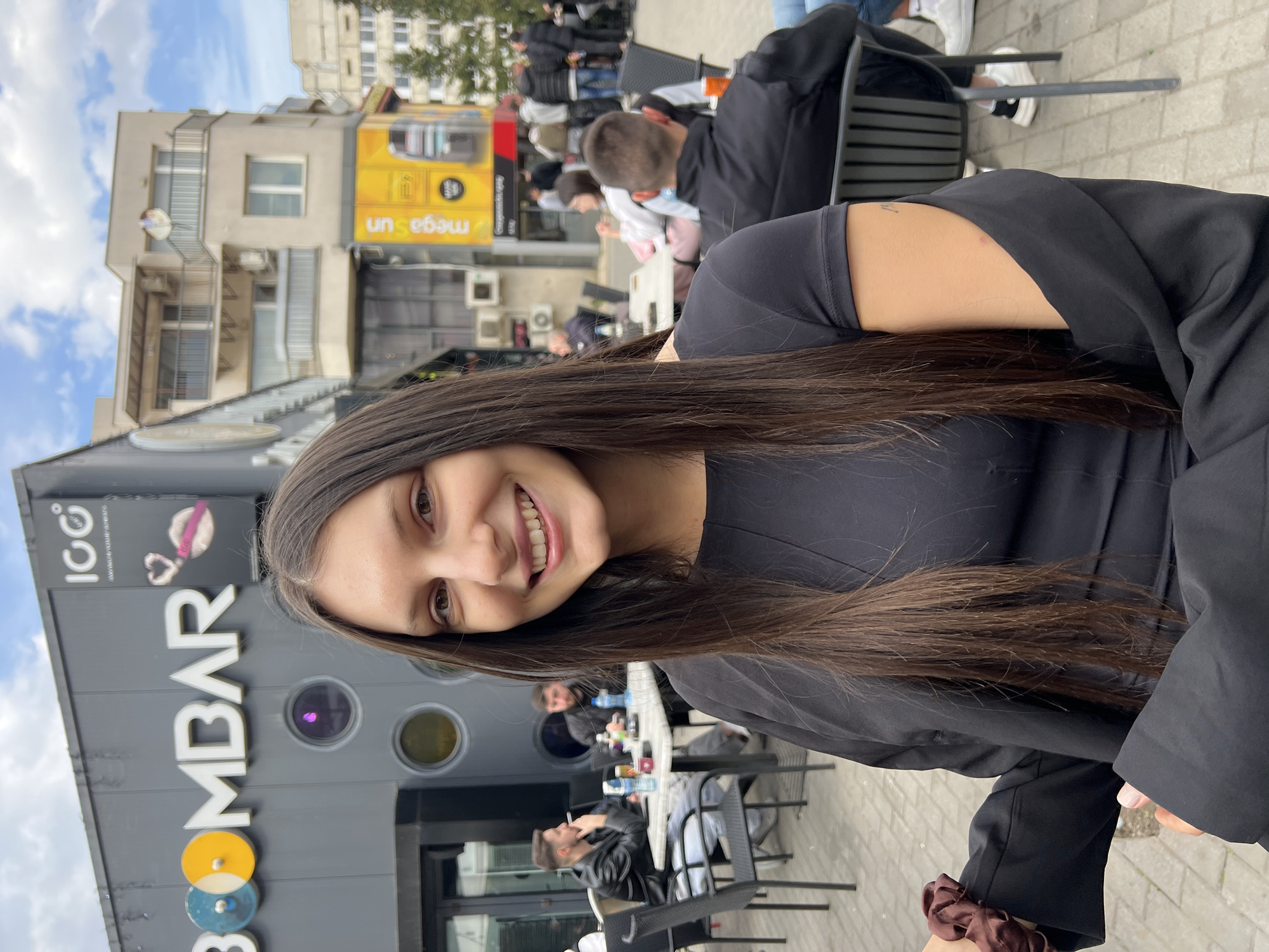 The image depicts a young adult female posing for the camera in an outdoor urban setting in Sofia, Bulgaria. The location is identifiable as an outdoor seating area adjacent to the "BOMBAR" establishment, situated at ul. "Knyaz Boris I" 175, 1000 Sofia Center.
The primary subject, positioned in the foreground and slightly right of center, is a female with long, straight dark brown hair, smiling directly towards the viewer. She is wearing a black short-sleeved top and a black long-sleeved jacket draped over her shoulders. Her left hand holds a brown scrunchie. A faint, small linear marking is visible on her right upper arm.
In the mid-ground, behind the main subject, several individuals are seated at black metal tables with matching chairs, part of the cafe's outdoor seating. To the left, a male wearing a black leather jacket and light-colored pants is seated at a table, looking left while holding what appears to be a lit cigarette. On his table are bottles and cups. Further back and to the right, another male is seen from behind, wearing a black vest or jacket, seated at a table with an orange cup. Other blurred figures populate the background, indicating a populated area.
The background features the exterior of the "BOMBAR" building, a dark gray structure with large white letters spelling "BOMBAR." Above "BOMBAR," a smaller sign displays "100" and "www.bombargroup.bg." To the right of the main sign, a pink graphic of lips with lipstick and a heart symbol is visible. The building has circular windows, some reflecting light. Adjacent to and behind "BOMBAR," a light-colored multi-story building with balconies and several outdoor air conditioning units is visible under a partly cloudy sky. To the far right, a yellow storefront with "mega sun" branding is prominent, with indistinct figures walking in front of it. The ground surface is paved with light-colored rectangular tiles.
