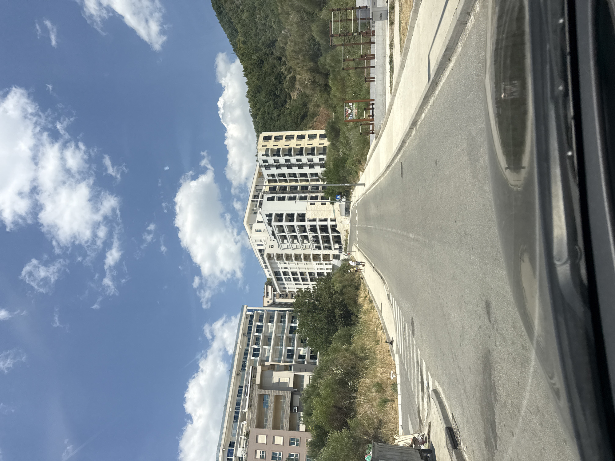 The image depicts a street scene in Boreti, Montenegro, on a clear day with scattered white clouds. In the foreground, the hood of a vehicle is visible, reflecting a portion of the surrounding environment. The street itself is paved and slopes upwards towards several modern apartment buildings. These buildings are multi-story structures with white facades and dark-railed balconies. Green trees and dense vegetation are situated between the street and the buildings, as well as on the hillside behind them. To the right of the street, there is a paved walkway and an outdoor exercise area with metal equipment. On the left side of the street, near a crosswalk, there is a large dumpster and some discarded items. Two individuals are walking on the sidewalk, heading uphill away from the viewer.