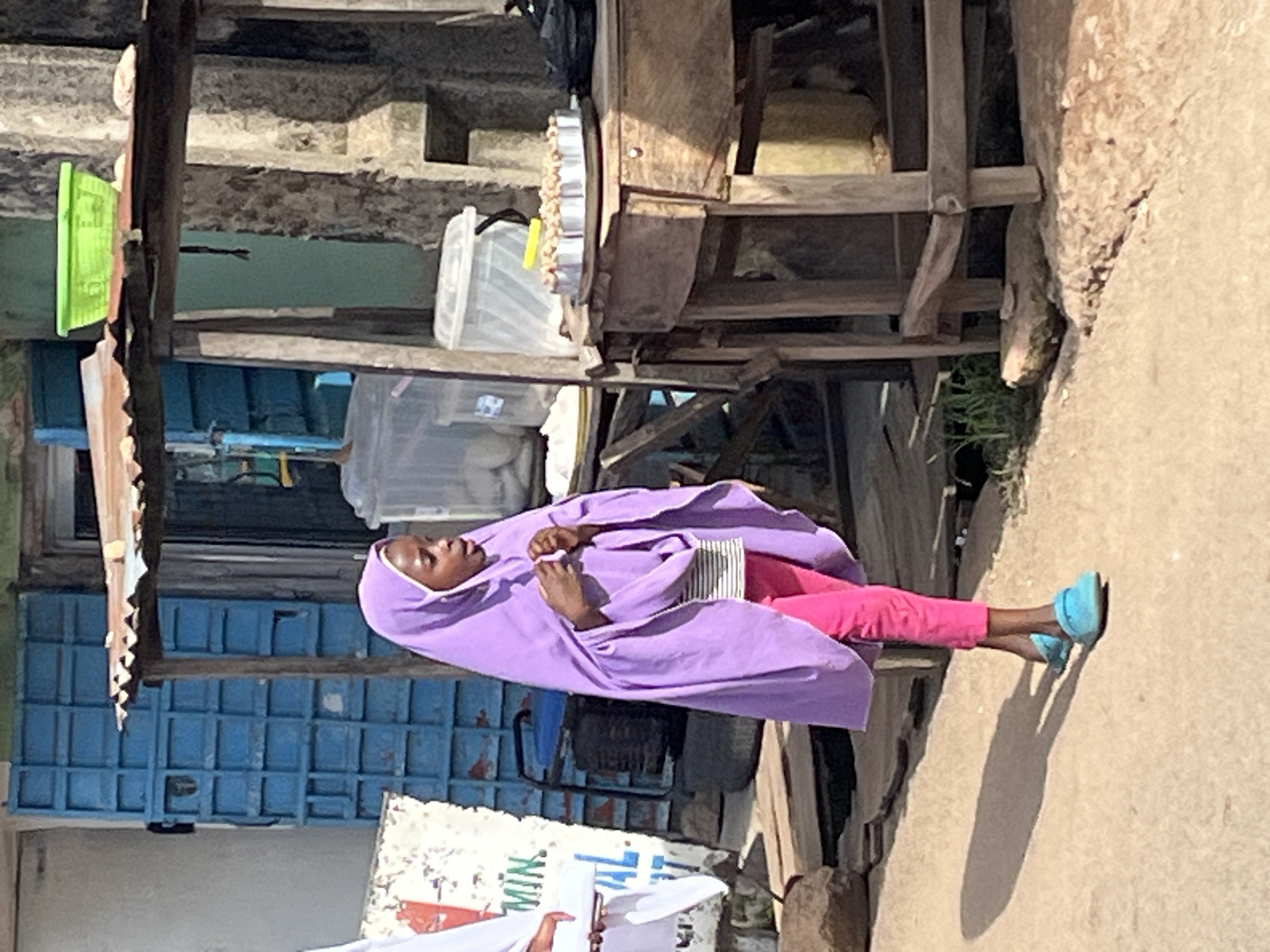 An outdoor scene in Jos, Nigeria, captures a young girl in a purple hijab and pink pants, walking on a dirt path. She is holding a piece of tissue paper. Behind her, a stall with a corrugated metal roof displays food items on a wooden table. To the left, a blue metal structure and part of a building are visible. The overall environment suggests a street-side market or commercial area.