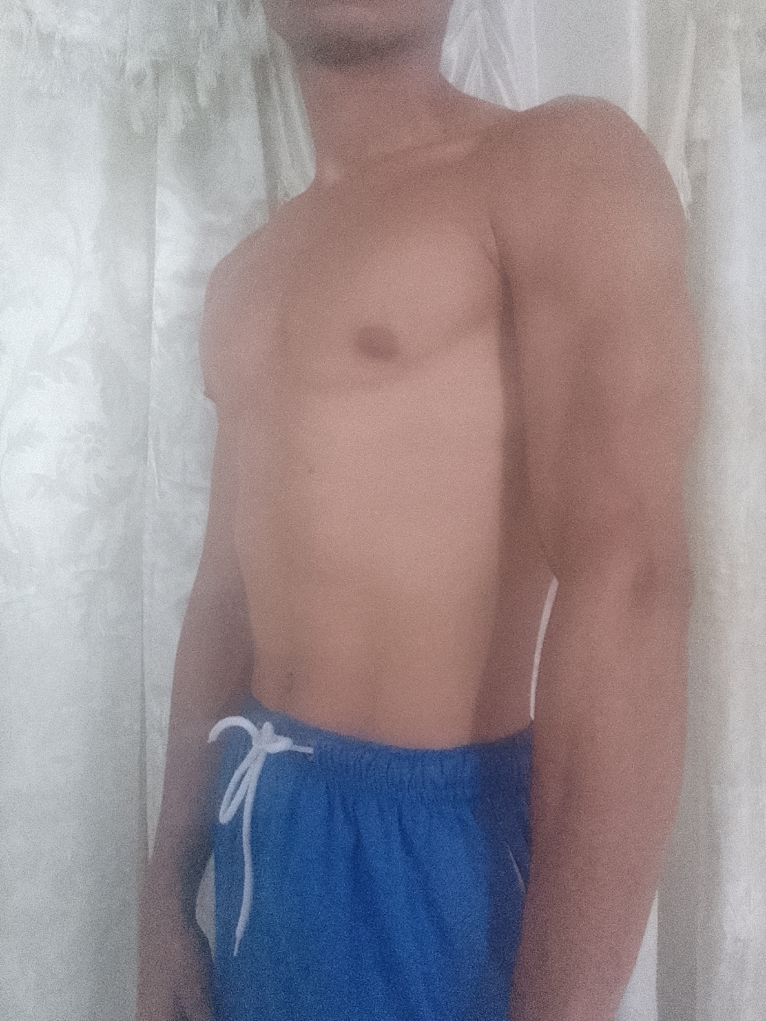 A shirtless individual wearing blue shorts with a white drawstring stands in front of a white curtain. The person's head is partially cropped out of the frame. No information suggests the location is Juan Griego, Venezuela.