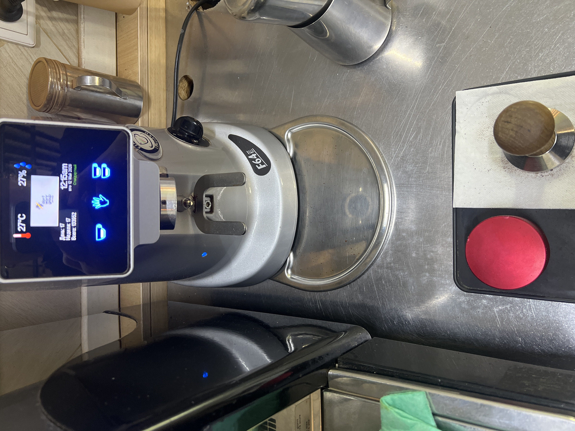 The image shows a close-up view of a coffee preparation station. The scene features a silver-colored coffee grinder with a digital display showing the time as 12:15 am, the date as May 12, 2026, and a temperature reading of 27°C. The grinder has a bean hopper and a dispensing mechanism. To the right of the grinder is a tamper and a red button, possibly for operating the espresso machine. The countertop appears to be stainless steel. The grinder is labeled "F64" and "ITALIANO." The image shows details indicating it might be in Russia, as the digital display includes Cyrillic characters. Given the availability of good coffee in the city, it could be reasonably inferred that the picture could have been taken in a cafe, restaurant, or someone's home in Saint Petersburg, Russia.