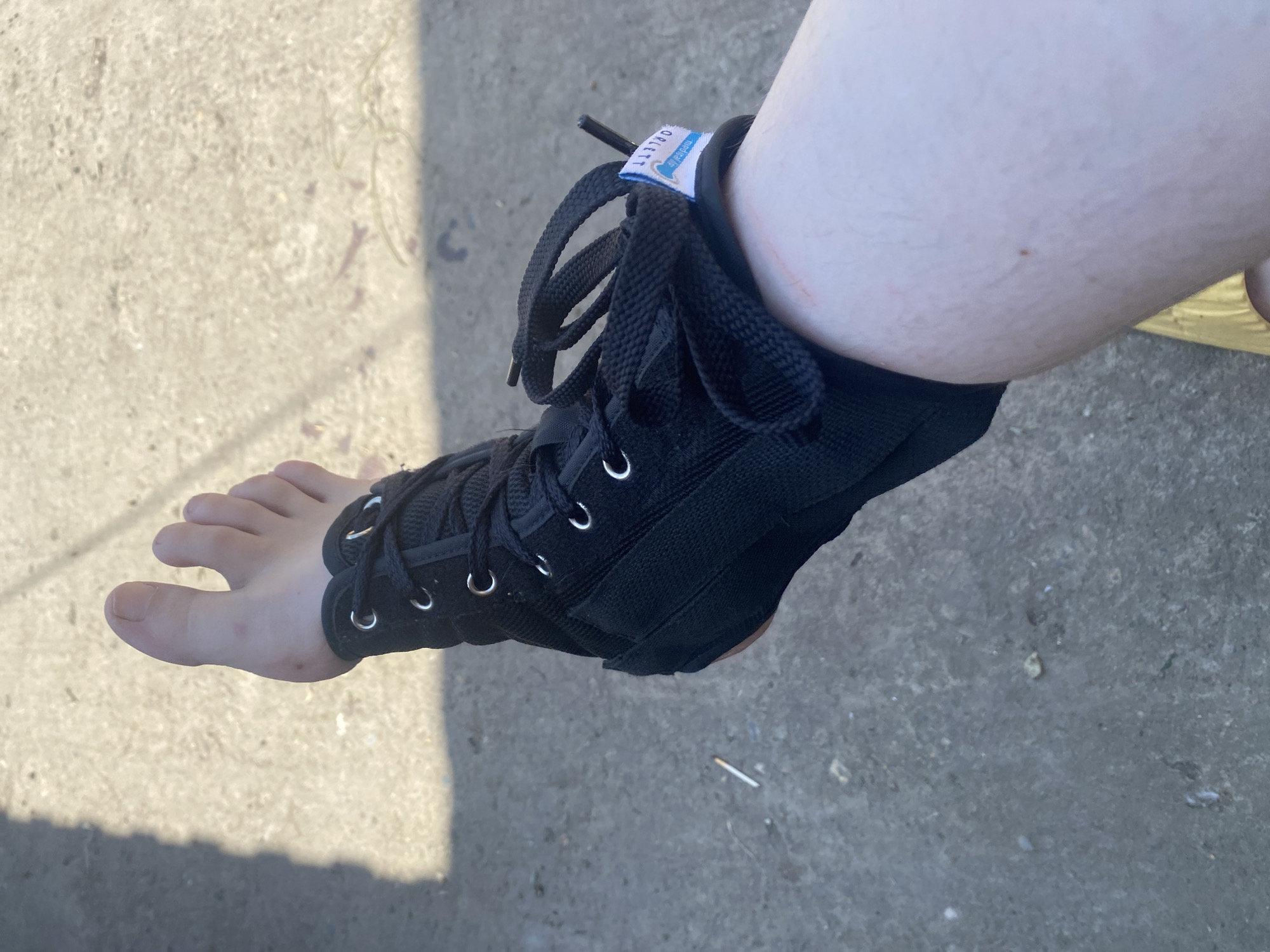 A fair-skinned human left lower leg and foot, wearing a black lace-up ankle brace, are depicted on a sunlit, uneven concrete surface in Kisliakovskaya, Russia. The foot is oriented with toes pointing left. The black fabric brace features silver-colored eyelets with black laces tied. A white and blue label, partially obscured and inverted, reads "OELLET" with "medical" text underneath, positioned on the outer side of the brace. The visible skin on the foot and lower leg shows minor blemishes, small moles, and fine hair. A slight red scratch is present on the skin immediately above the brace on the ankle. The concrete ground exhibits scattered small debris and a distinct, elongated shadow extends across the upper-left quadrant. A single discarded cigarette butt is visible on the concrete in the mid-left area of the frame.