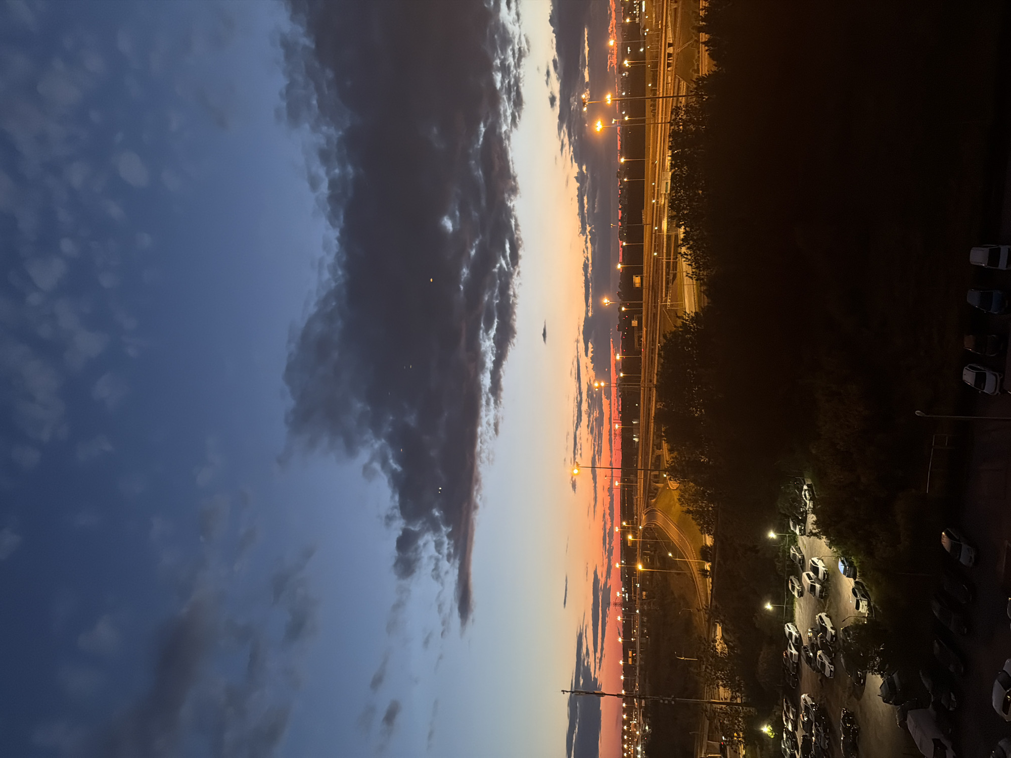The image depicts an urban landscape at dusk or dawn. The sky is a gradient of deep blue to fiery orange, with dark, heavy clouds obscuring parts of the view. Several streetlights illuminate a complex network of highways and roads, casting a warm glow on the asphalt. In the foreground, a parking lot is filled with numerous cars. Some cars are parked in neat rows, while others appear to be scattered. The scene is dominated by the transition from daylight to darkness, with artificial lights taking over as the primary source of illumination. The overall impression is one of a bustling urban environment at the cusp of night.
