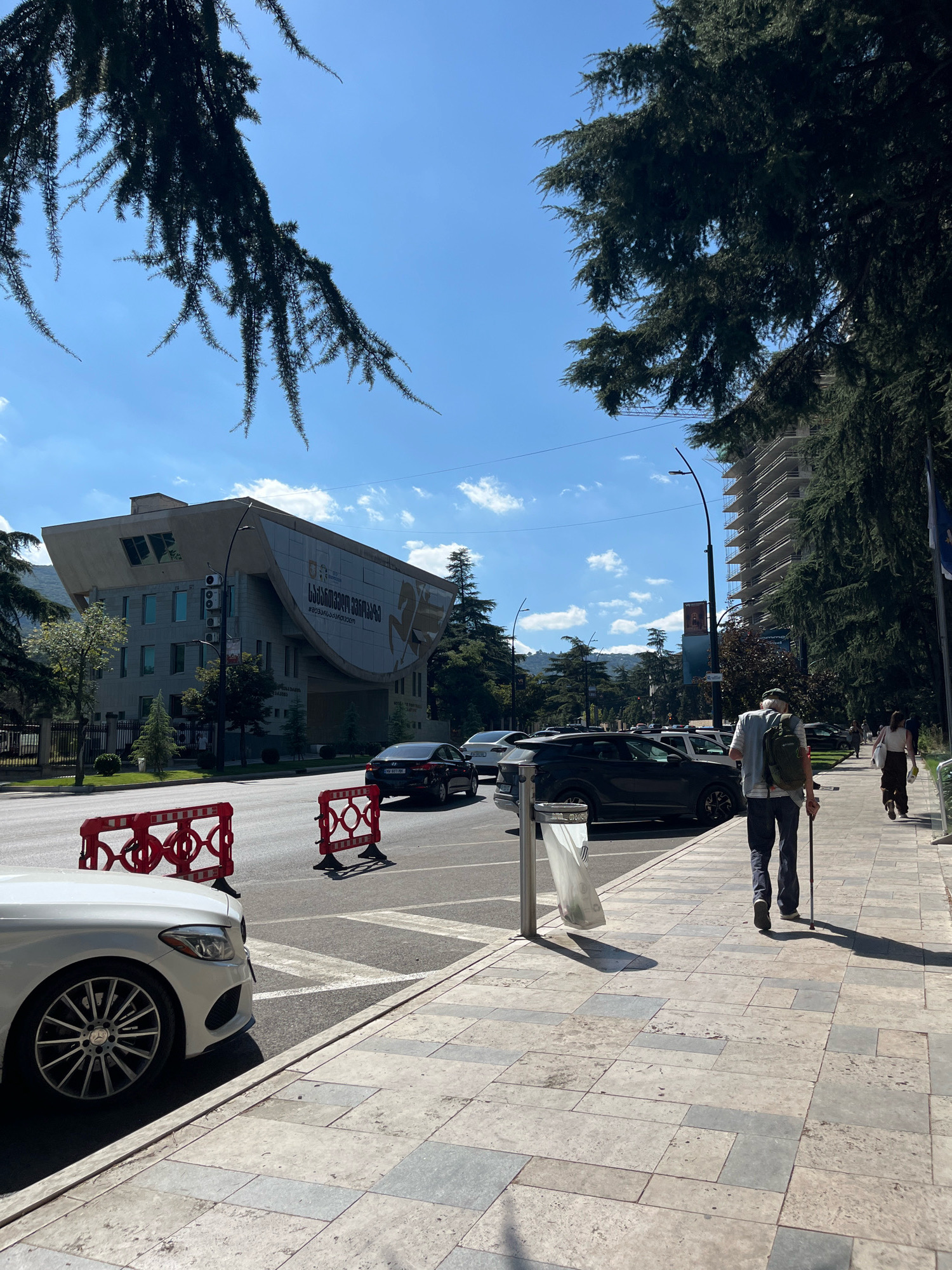 The image depicts a street scene in Tbilisi, Georgia, likely in an urban area with a mix of modern architecture and established trees. In the foreground, a white Mercedes-Benz is parked along the curb, its front wheel and fender visible. Red plastic barriers are placed on the road, suggesting temporary construction or traffic diversion. A sidewalk runs alongside the street, paved with large, light-colored tiles.
On the sidewalk, a man wearing a backpack and a cap walks away from the camera, using a walking stick. Another pedestrian, a woman, is further ahead on the sidewalk. Several cars are visible on the road, including a black SUV and a dark sedan.
The background features a distinctive modern building with a curved roof and large graphic elements on its facade. To the right, a tall, unfinished building under construction is visible, surrounded by scaffolding. The environment is characterized by lush green trees lining the street, casting shadows on the sidewalk. The sky is bright blue with scattered clouds, indicating a sunny day. The scene appears to be a typical urban setting in Tbilisi during daylight hours.