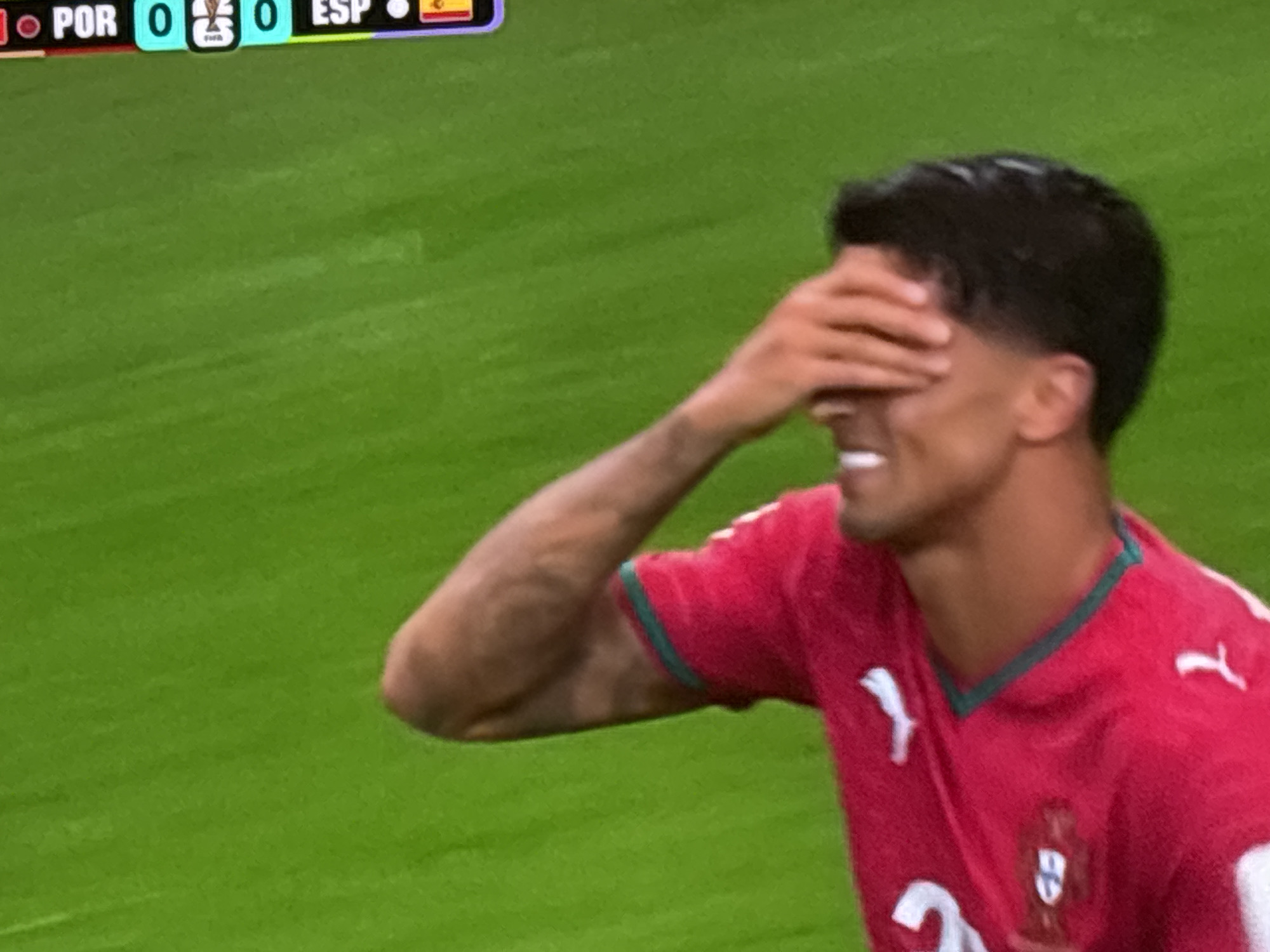 The image is a close-up shot of a male soccer player on a bright green field. He is wearing a red jersey with a green trim around the collar and white detailing. His right hand is covering his eyes, and he appears to be smiling or grimacing. The player's skin tone is medium, and his hair is dark and short. In the background, a scoreboard shows "POR 0 0 ESP" with flags indicating Portugal and Spain, along with the FIFA World Cup trophy icon. The background is out of focus, showing the expanse of the field. The overall impression is of a player reacting to a moment in a soccer match. There is no indication of the city of Burgas, Bulgaria, being present in the image.