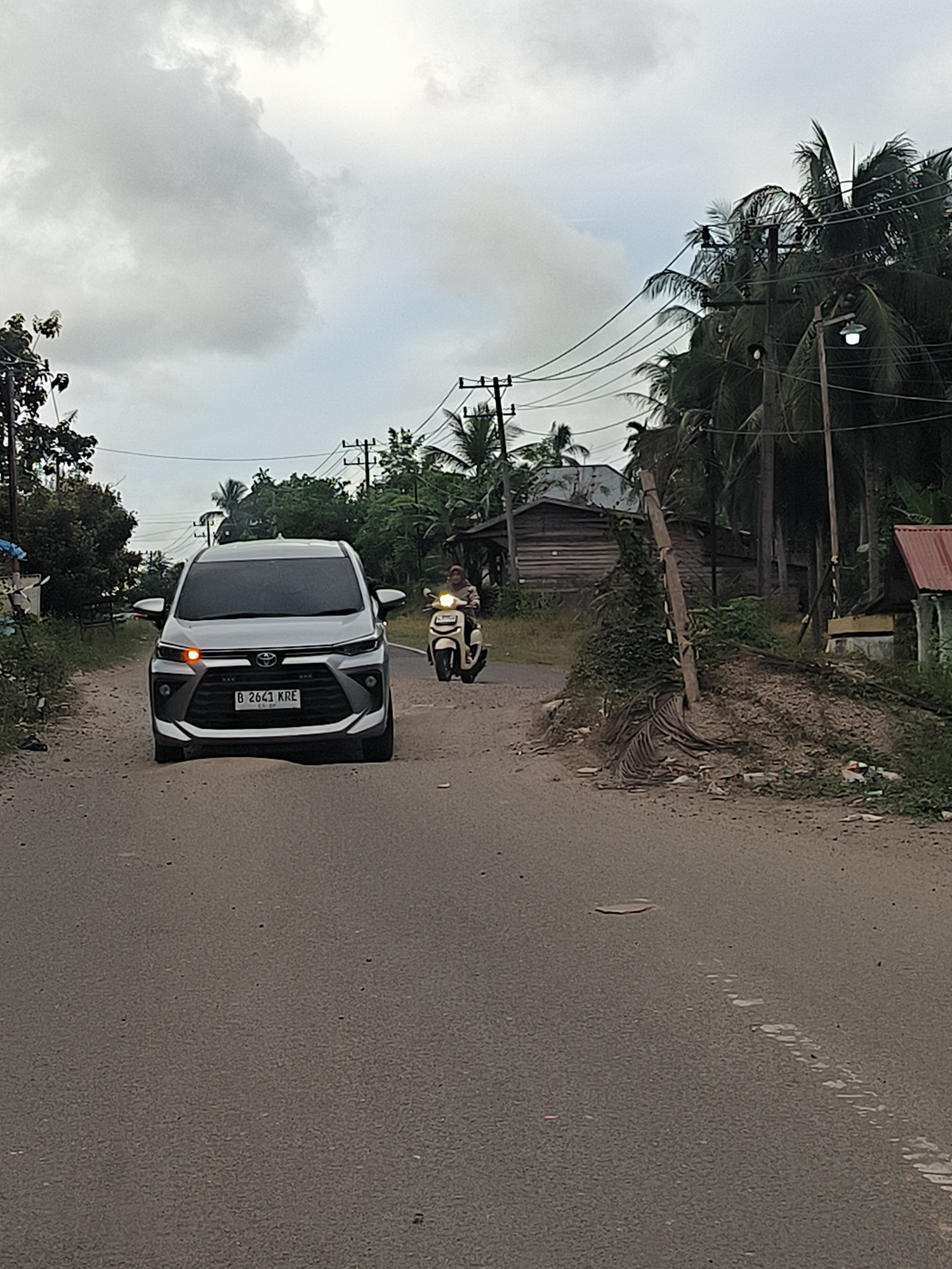 A silver Toyota Avanza, license plate B 2641 KRE, is parked on a dirt shoulder next to a paved road in Payadua, Indonesia. The car's hazard lights are on. To the right of the car, a person on a light-colored scooter is driving away from the camera. The environment is rural, with palm trees, wooden buildings, and utility poles visible along the roadside. The sky is overcast, suggesting a cloudy day.