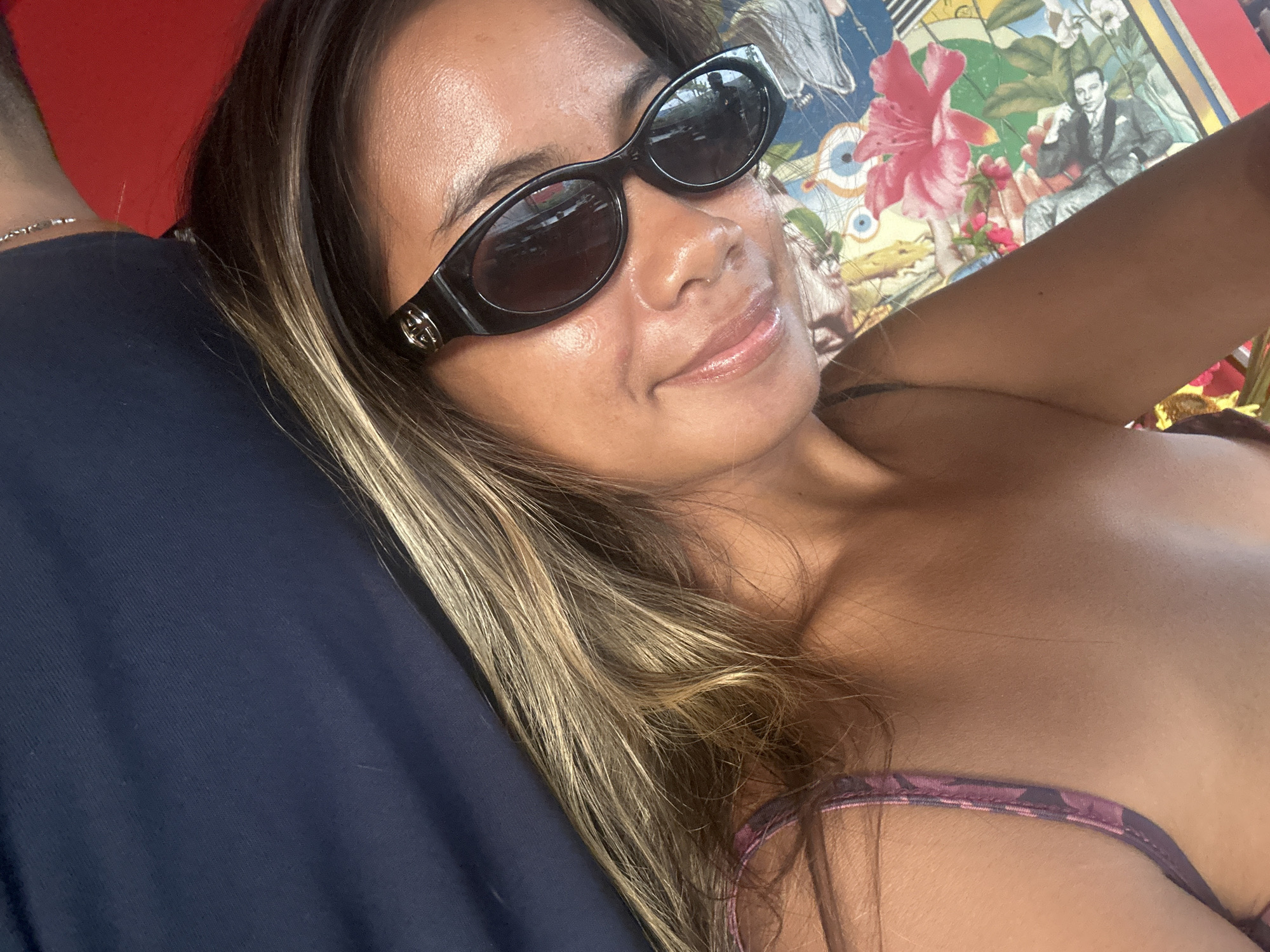 A close-up, high-angle selfie shows a woman with long, wavy brown and blonde hair wearing black, cat-eye sunglasses with a gold logo. She is looking towards the camera with a slight smile. She is wearing a purple and black floral patterned top. The background is a colorful, collage-like pattern with images of flowers and faces. The woman is lying down, and a dark blue fabric is visible to her left, along with a red surface to her right. The image does not provide enough information to determine the location within North Kuta, Indonesia.