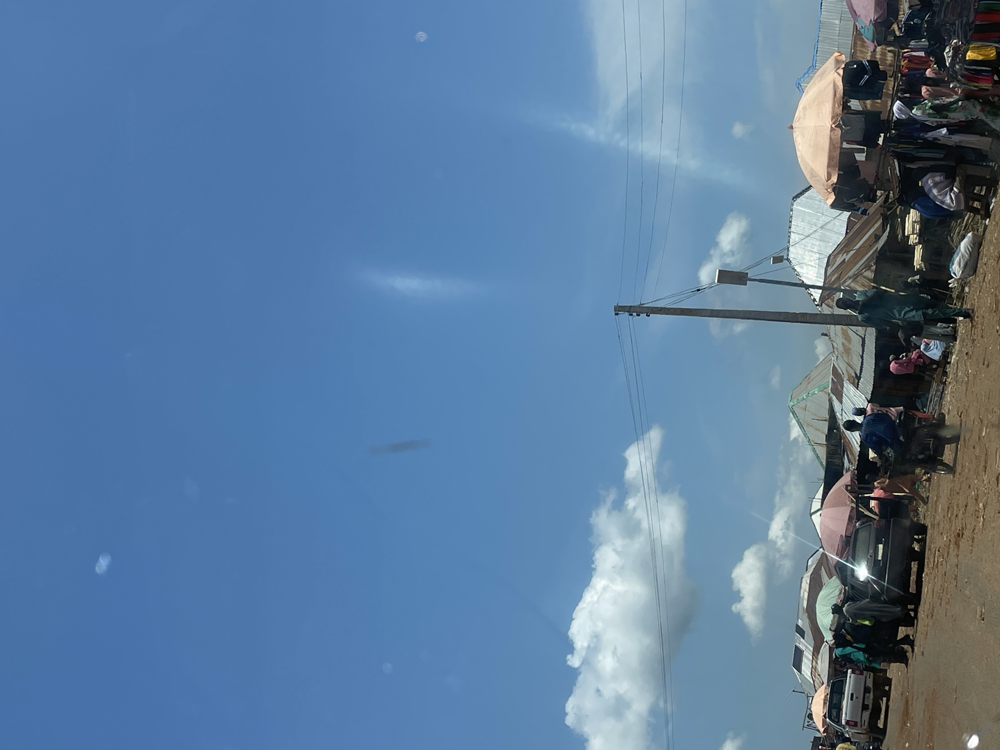 This is a street-level photograph of a bustling market scene in Zalau, Nigeria. The foreground features a dirt road with several vehicles, including a white pickup truck, a dark sedan, and a motorcycle. People are milling about the vehicles and stalls. Several market stalls are set up, some covered by umbrellas and awnings. Various goods are displayed, including clothing hanging from racks. The background shows a cluster of buildings with corrugated metal roofs. The sky above is bright blue with scattered clouds. A power line supported by a utility pole runs across the upper portion of the scene. The overall impression is one of a vibrant and active marketplace under a clear sky.