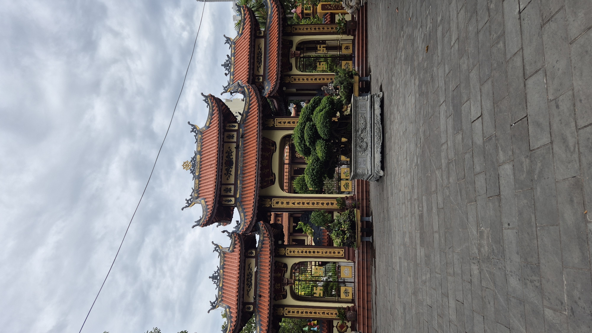 The image depicts the entrance to a Buddhist temple, possibly Chua Tam Thai, located in the city of Đà Nẵng, Vietnam. The architecture features traditional East Asian temple design with multiple tiered, curved roofs adorned with red tiles and decorative accents. The main gate is a grand structure with ornate carvings and calligraphy, flanked by two smaller, similar structures. In front of the gate, a large, well-manicured bonsai tree sits in a decorative stone planter, adding a touch of nature to the scene. The ground is paved with large, rectangular stone tiles. The sky is overcast with a cloudy, grey appearance, suggesting a cool or humid day. The overall impression is one of serene architectural beauty and cultural significance.