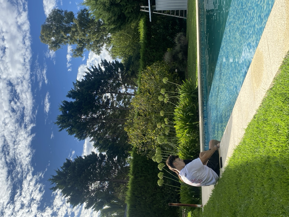 A person sits at the edge of a pool, facing away from the camera, dressed in a white shirt and dark shorts. The pool's edge is a light tan color and the pool itself is surrounded by lush green grass and various plants. Tall green trees tower in the background against a bright blue sky filled with fluffy white clouds.