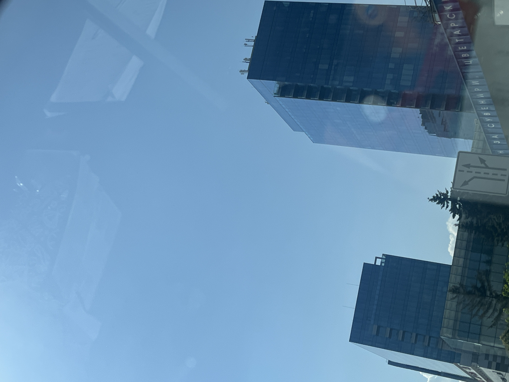 A daytime, low-angle outdoor shot captures a clear blue sky with a few faint reflections. Two modern, dark glass skyscrapers dominate the view, one taller and angled to the right, the other to the left. The taller building shows exposed concrete on its side, indicating it may be under construction or undergoing renovation. Along the bottom of the frame, a reflective sign with Cyrillic lettering, "Н ЗА СУВЕНИРИ ЦВЕТАРСКИ," is visible. A road sign with directional arrows for three lanes is also present to the left of the sign. Several evergreen trees are interspersed between the buildings and in the foreground.