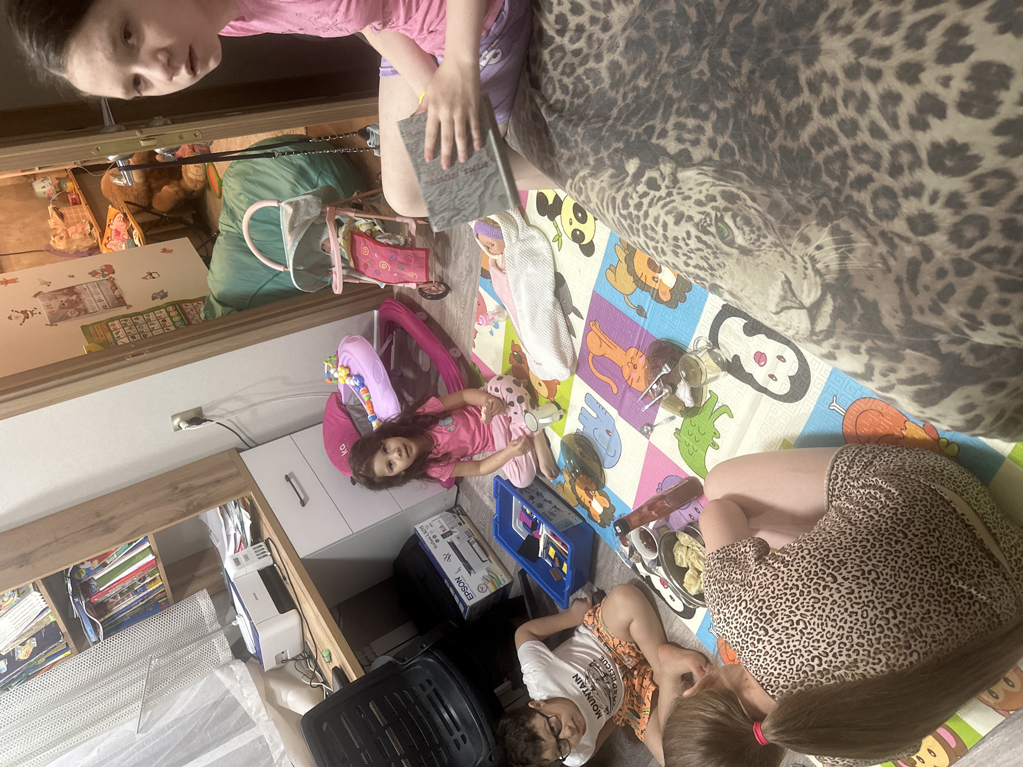 The image captures an indoor scene where several individuals, likely children, are interacting within a domestic setting. The environment suggests a child-friendly space with an animal-themed blanket, play mats adorned with animal illustrations, and toys scattered around. One individual, seen from an elevated perspective, is holding a book. A girl, wearing pink clothing, is lying on the play mat, seemingly engaged in play. Another child, dressed in a t-shirt and shorts, is also lying on the floor, close to a tray containing food. A woman is positioned towards the bottom right corner of the image, her upper body angled towards the children. Background elements include a shelf filled with books, a printer, and other miscellaneous objects.