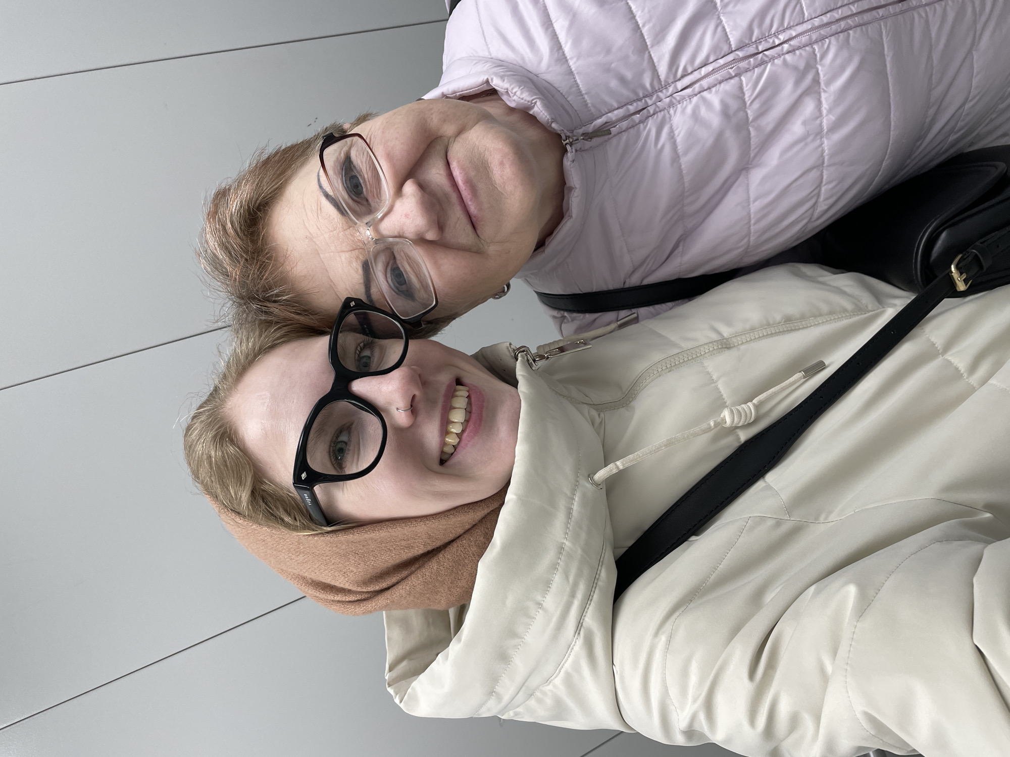 Two women are posing closely together for a selfie. The woman in the foreground is younger, with blonde hair, wearing black framed glasses, a tan scarf, and a white jacket with a black strap across her torso. She is smiling with a visible nose ring. The woman behind her is older, with graying hair, wearing glasses with a brown and black frame, and a light purple quilted jacket.
The background is a light gray, slightly textured surface. There is no specific information to place the location in the city of Izhevsk, Russia.