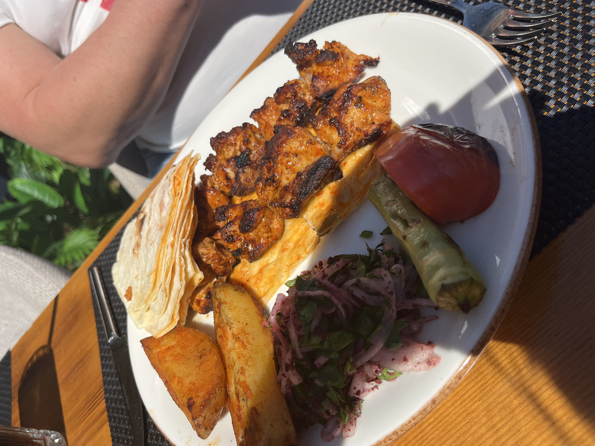 A high-angle shot showcases a white, oval-shaped plate brimming with food. The main element of the meal appears to be grilled chicken kebab pieces arranged on top of a thin flatbread. Accompanying the kebab are roasted potato wedges, grilled tomato, roasted green pepper, and a salad of onions and herbs. There are some thin flat bread folds towards the left. Silverware is seen positioned on the left and upper right corner of the plate. The plate rests on a wooden surface with a black woven texture. Part of an arm and a white shirt are visible in the upper left corner. The location is possibly in Budva, Montenegro.