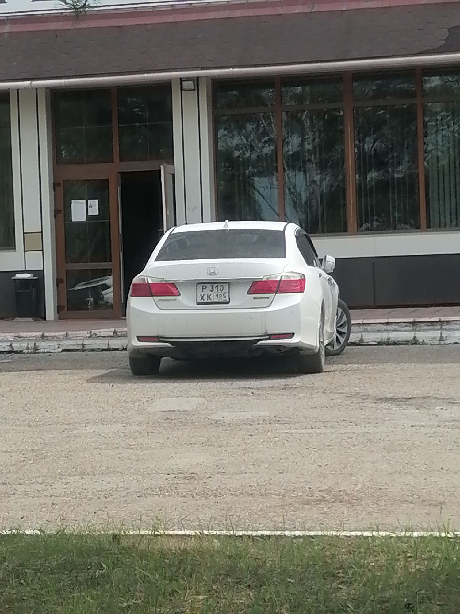 A white sedan is parked on a gravel lot in front of a building with large windows and a dark-tiled roof. The car is facing away from the camera, and its license plate is visible. The license plate reads "P 310 XK 125". The building appears to be a commercial or public building. The environment is outdoors, with some dry grass in the foreground. The location is the city of Lesozavodsk, Russia.