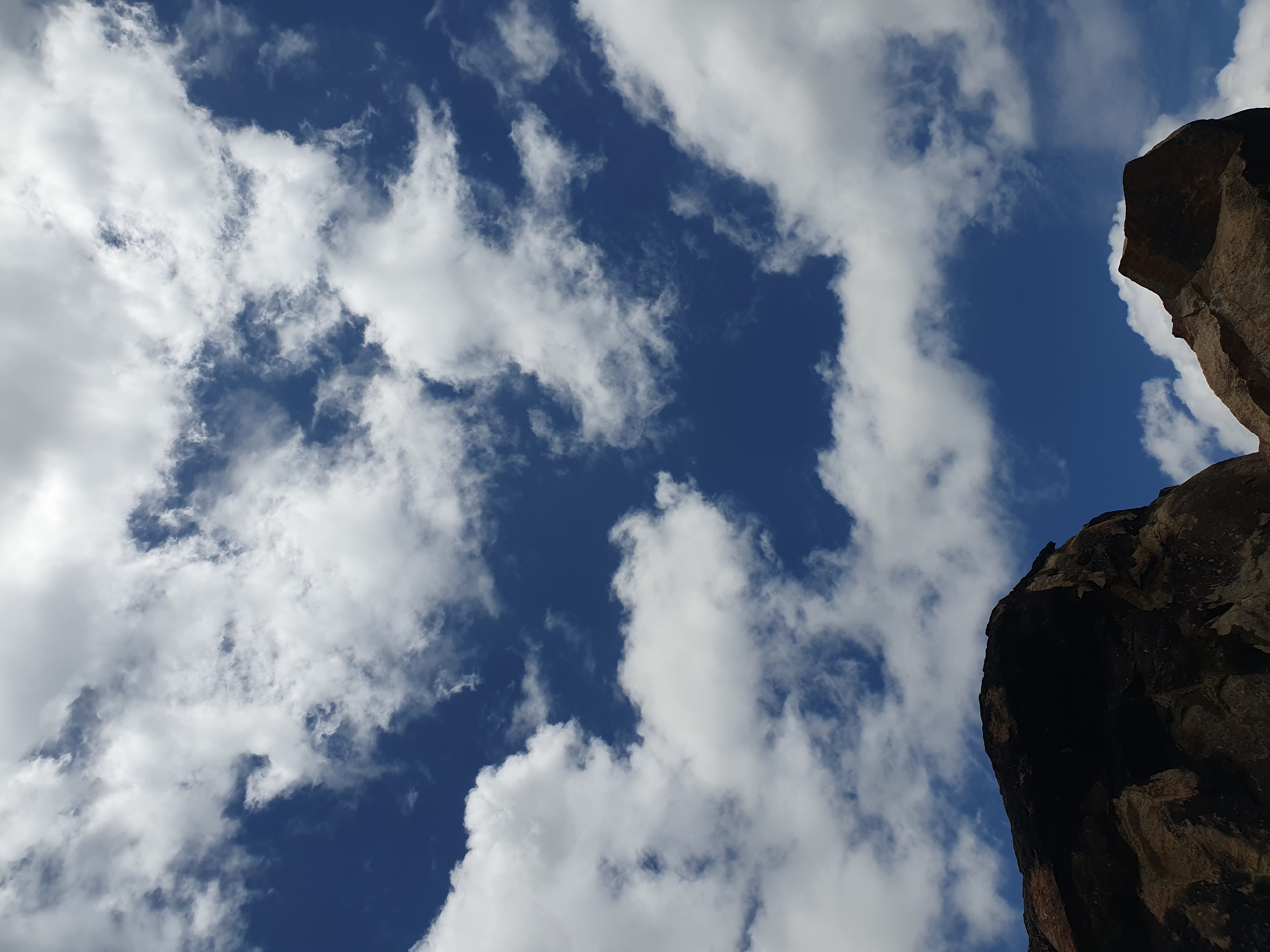The image depicts a striking view of the sky from the perspective of Gura Top, Nigeria. The deep blue sky is adorned with fluffy white clouds, creating a dynamic contrast. In the foreground, the dark silhouettes of rocky formations frame the lower portion of the image. These rocks appear rugged and textured, with hints of earthy tones visible where light catches their surfaces. The composition emphasizes the vastness of the sky against the grounding presence of the natural rock formations, suggesting an outdoor landscape in a mountainous or elevated area. There are no people or visible actions taking place in the scene.
