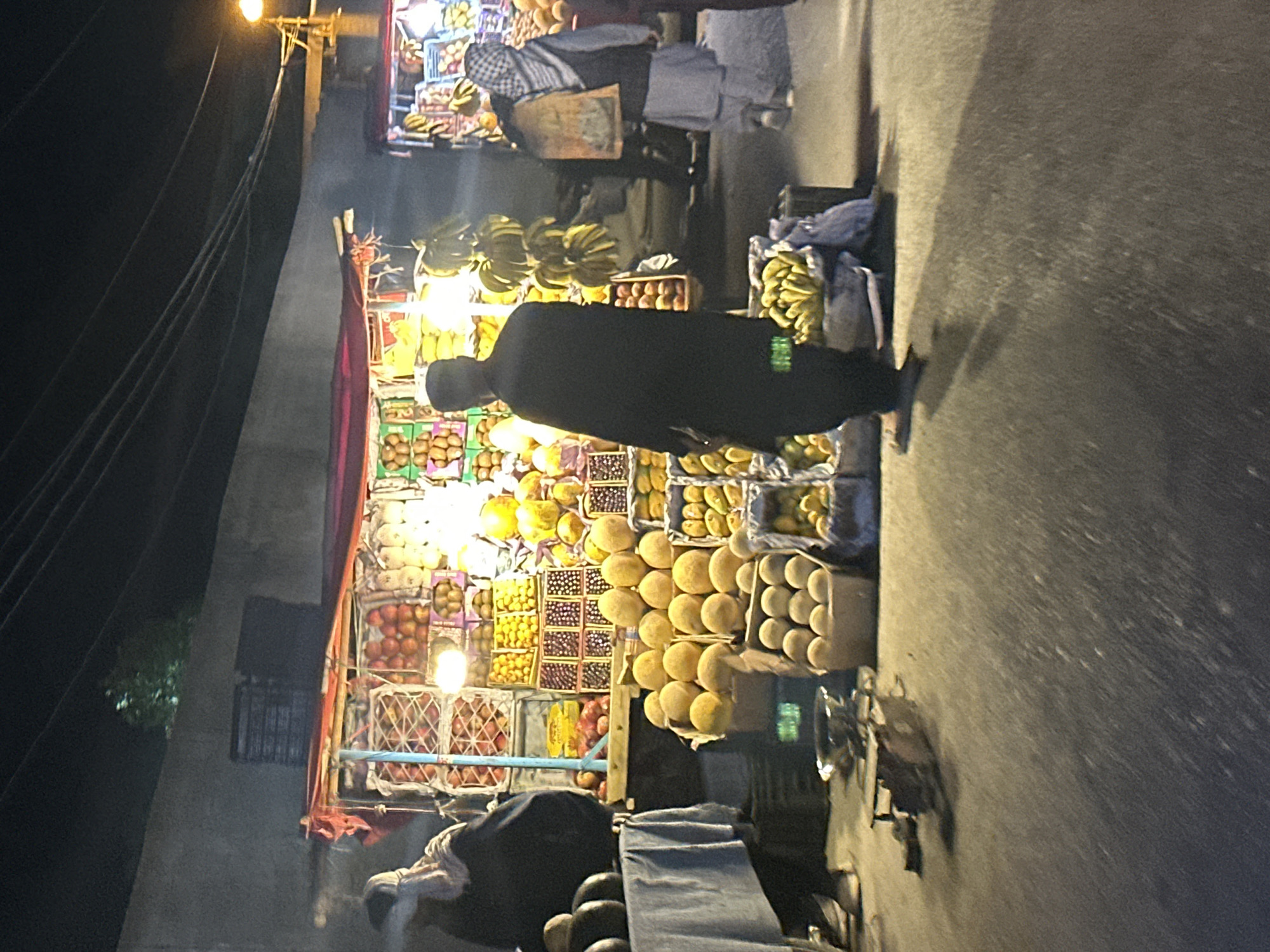 A nighttime street scene in Quetta City Tehsil, Pakistan, shows a brightly lit fruit stand overflowing with various fruits. A man in a dark robe and cap stands near the stand, possibly a customer. Another person is behind the fruit stand, and people are walking past. The street is dark and paved. Wires run across the left side of the image.