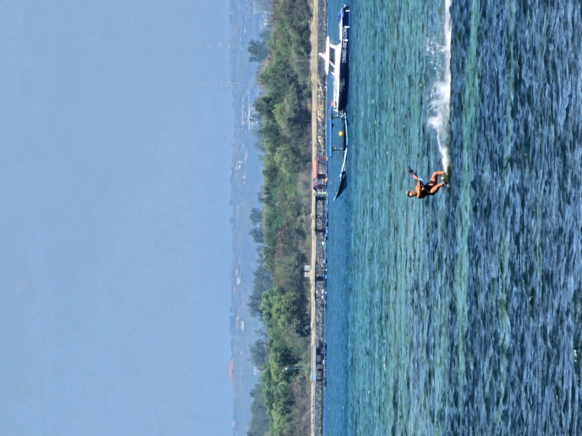 A woman kiteboards on a bright, sunny day in the ocean off Denpasar, Indonesia. She is wearing a black swimsuit and a helmet and is in motion, leaving a spray of water behind her. The water is a vibrant turquoise and appears shallow in places, with visible ripples and waves. In the middle ground, there is a long pier with structures and aquaculture cages. Further back, a white boat with a blue stripe and "DIVING-INDONESIA.COM" written on its side is moored near a smaller traditional boat. The background features a lush, green coastline with trees and some buildings on a gentle slope, leading to a hazy, pale blue sky. The overall scene depicts a recreational activity in a coastal environment.