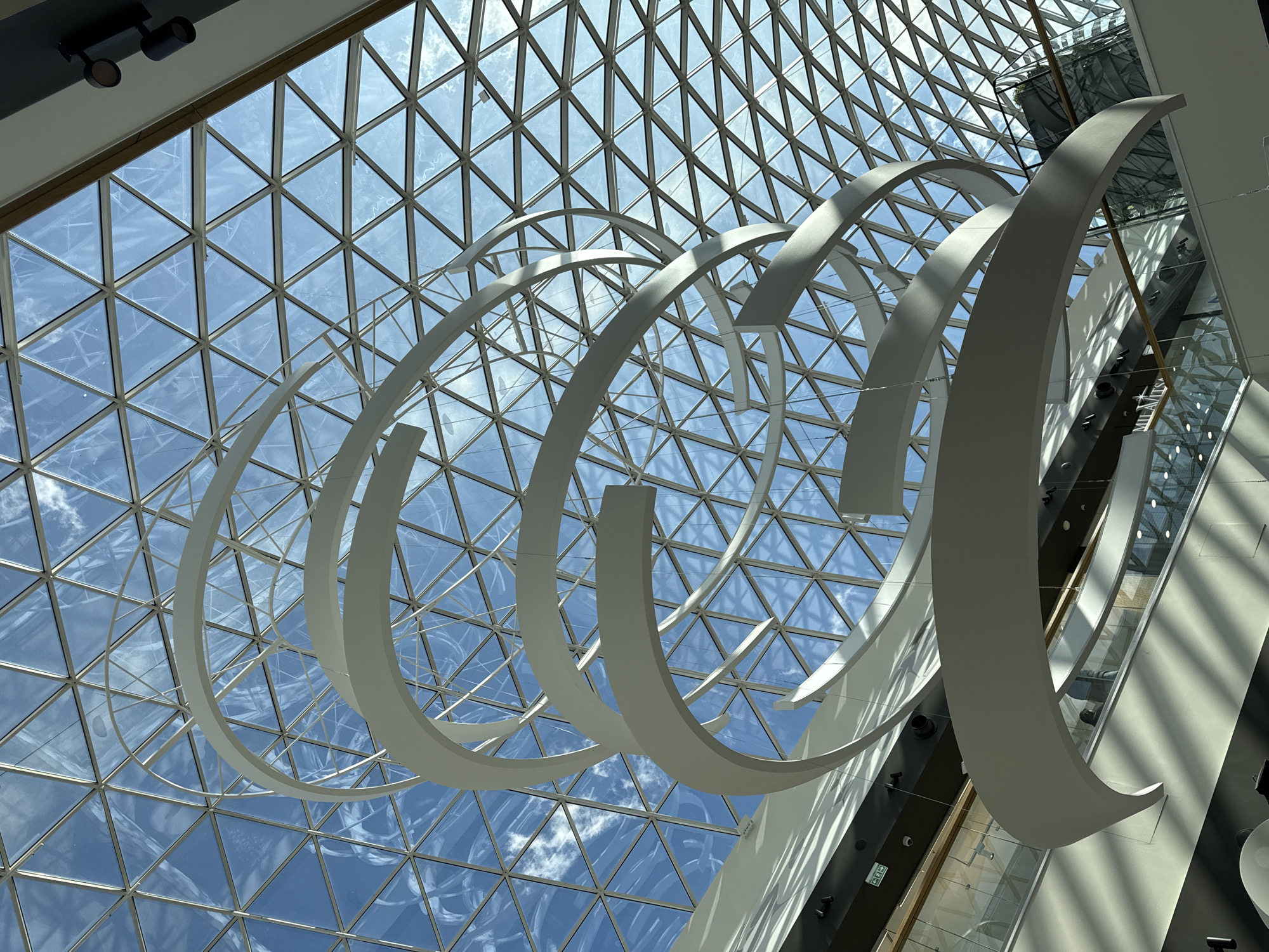 The image captures the interior of a large, modern building, looking upwards towards a vast, geometric glass ceiling that allows abundant natural light to flood the space. The ceiling is composed of numerous triangular glass panels set within a structural grid, resembling a geodesic dome or a similar architectural design. Through the clear glass, a bright blue sky with scattered white clouds is visible, indicating daytime.
Suspended from this ceiling are multiple large, white, curvilinear sculptural elements that coil downwards in an abstract, helical or spiral pattern. These art installations or architectural features cast distinct shadows on themselves and the building's interior surfaces due to the bright overhead sunlight.
The setting appears to be a spacious atrium or a similar public area within a contemporary building, possibly a shopping mall, convention center, or museum, located in Perm, Russia. No people are visible, giving the scene a serene and architectural focus.
Other visible architectural elements include dark and light-colored walls, possibly glass and metal railings or partitions for an escalator or elevator shaft, and recessed light fixtures along the edges of the ceiling or upper walls. A small green "EXIT" sign with white text and a running man symbol is discernible in one of the lower-right sections of the building's structure. The overall impression is one of grand scale, modern design, and brightness, emphasized by the interplay of natural light and shadow on the structural and sculptural forms.
