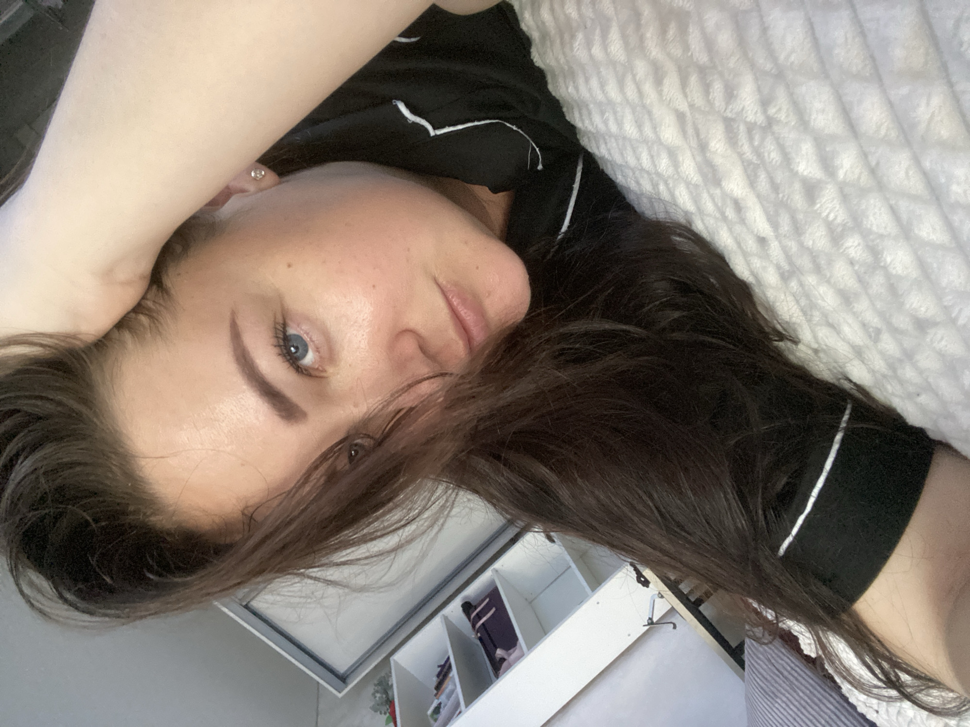A woman is lying on a white quilted bed. She is wearing black pajamas with white piping. Her dark hair is partially covering her face. She has blue eyes. Her left arm is raised above her head. In the background, there is a white shelf unit with various items. The location of the image cannot be definitively determined as Sochi, Russia, based solely on the visual information provided.
