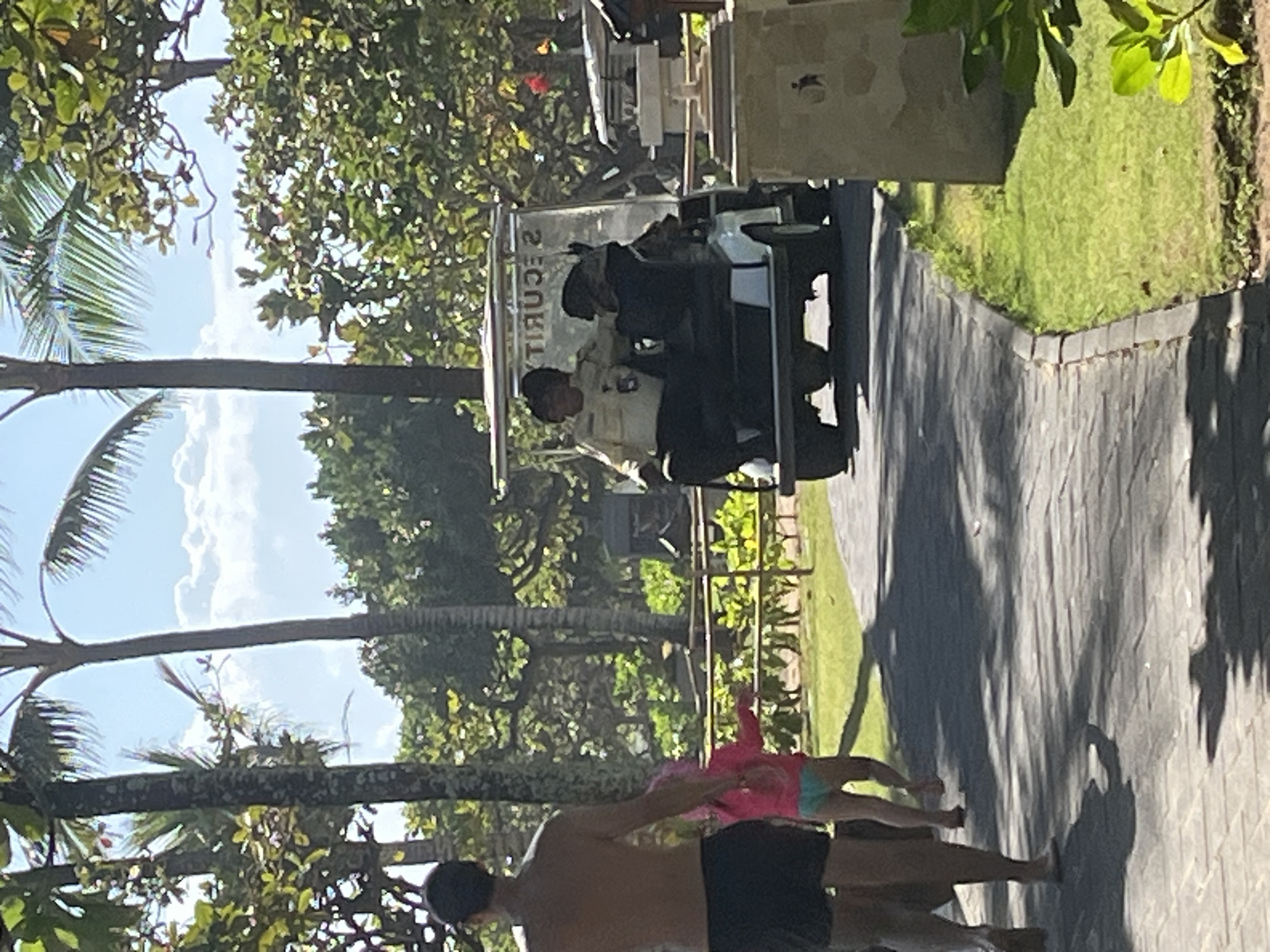 In Kuta Selatan, Indonesia, on a paved pathway, a person walks away from the camera, wearing dark shorts. A child in a pink top and blue bottoms walks ahead of them. Behind them, a white golf cart is driving on the pathway. Two individuals are seated in the golf cart; the driver wears a light-colored shirt and a dark cap, and the passenger wears dark clothing. The golf cart has "SECURITY" written on its windshield. The environment is tropical, with palm trees and lush green foliage. The sky is bright blue with scattered clouds.