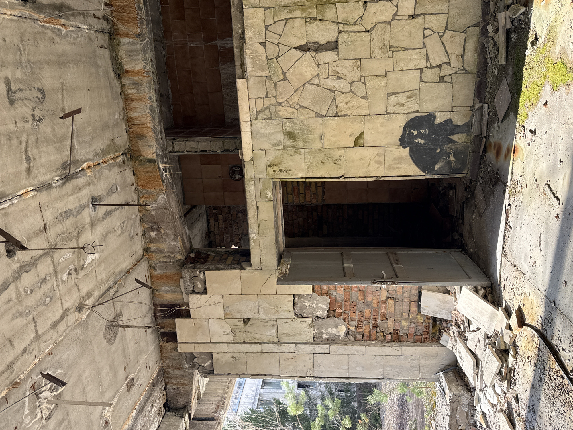 The image depicts the interior of an abandoned building in Pripyat, Ukraine. The building exhibits significant decay, with crumbling walls, exposed brick, and debris scattered across the floor. A doorway is visible, opening into a dark interior space. A dark graffiti mark is on the wall near the doorway. The environment is clearly post-apocalyptic, suggesting the aftermath of a disaster or abandonment. No people or subjects are directly visible.