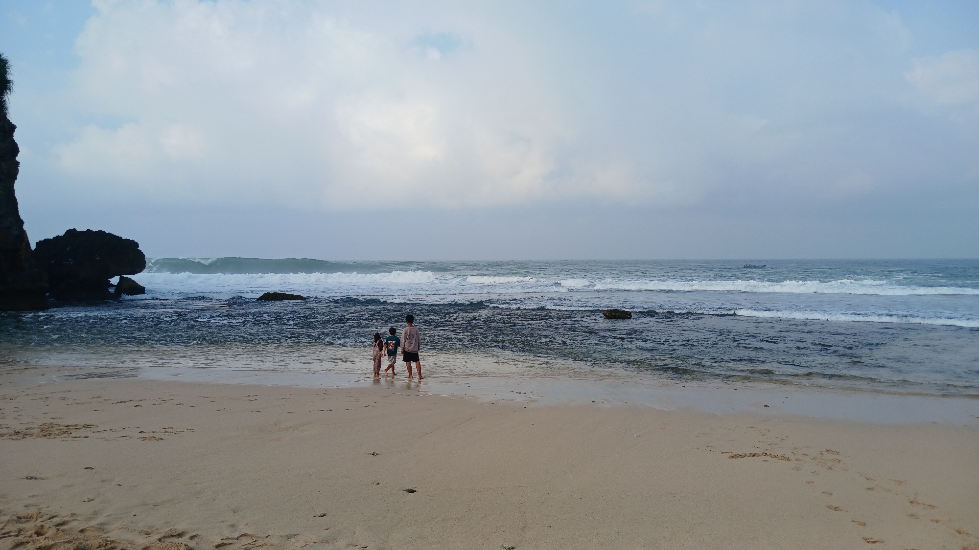 Three children stand on a sandy beach in Tanjungsari, Indonesia, facing the ocean. The sky above is overcast with white and grey clouds. Waves crash on the dark, rocky shore to the left, and further out, a small boat is visible on the water. The sand is damp near the water's edge, with footprints leading across it. The overall atmosphere is serene and natural.