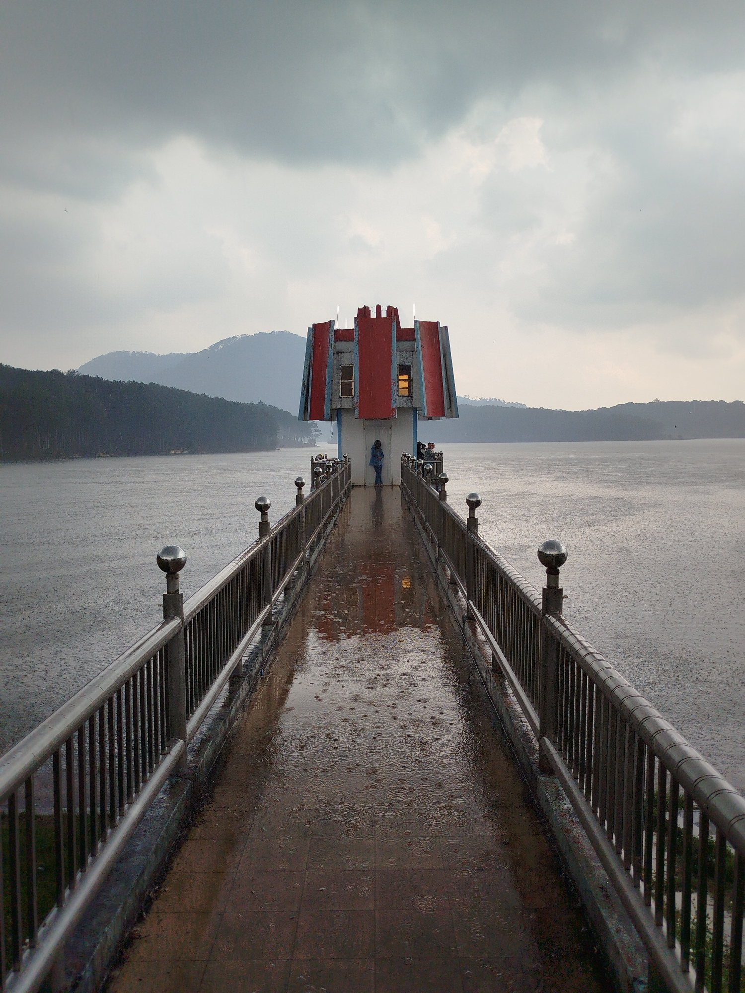 The image captures a gray, rainy day scene at Xuan Huong Lake in Da Lat, Vietnam. A narrow pedestrian bridge with metal railings extends into the lake toward a small building with a red-roofed structure on top. At the end of the bridge at the structure are some people. Rain falls heavily, creating ripples on the lake surface and a wet, reflective surface on the bridge. Hills and trees are visible in the background across the lake, shrouded in mist. The sky is overcast with gray clouds.