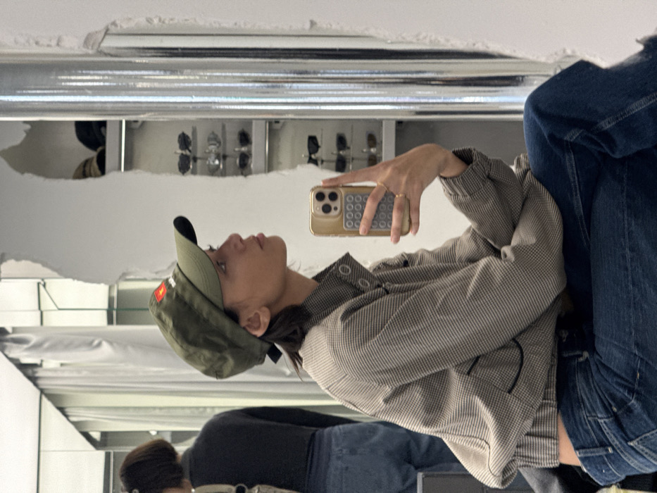 A young woman is depicted in a mirror selfie, captured in profile from the chest to the mid-thigh, looking towards the right side of the frame. She wears an olive green baseball cap with a red and white emblem on the front, a light brown and white patterned bomber jacket with a high collar and silver-colored snaps, and blue denim jeans. Her left hand holds a gold-colored smartphone, equipped with a light gray circular-patterned accessory, positioned to capture the reflection. A simple gold-colored ring is visible on her left ring finger. The environment is an indoor retail or fitting room setting within Paris, France, characterized by large reflective surfaces and white walls. A silver-colored vertical column is present on the right, alongside shelving displaying multiple pairs of sunglasses and other items, visible through a rough-edged opening in the background. A partial reflection of another individual, seen from behind, wearing a dark top and blue jeans, is visible in the upper left background.