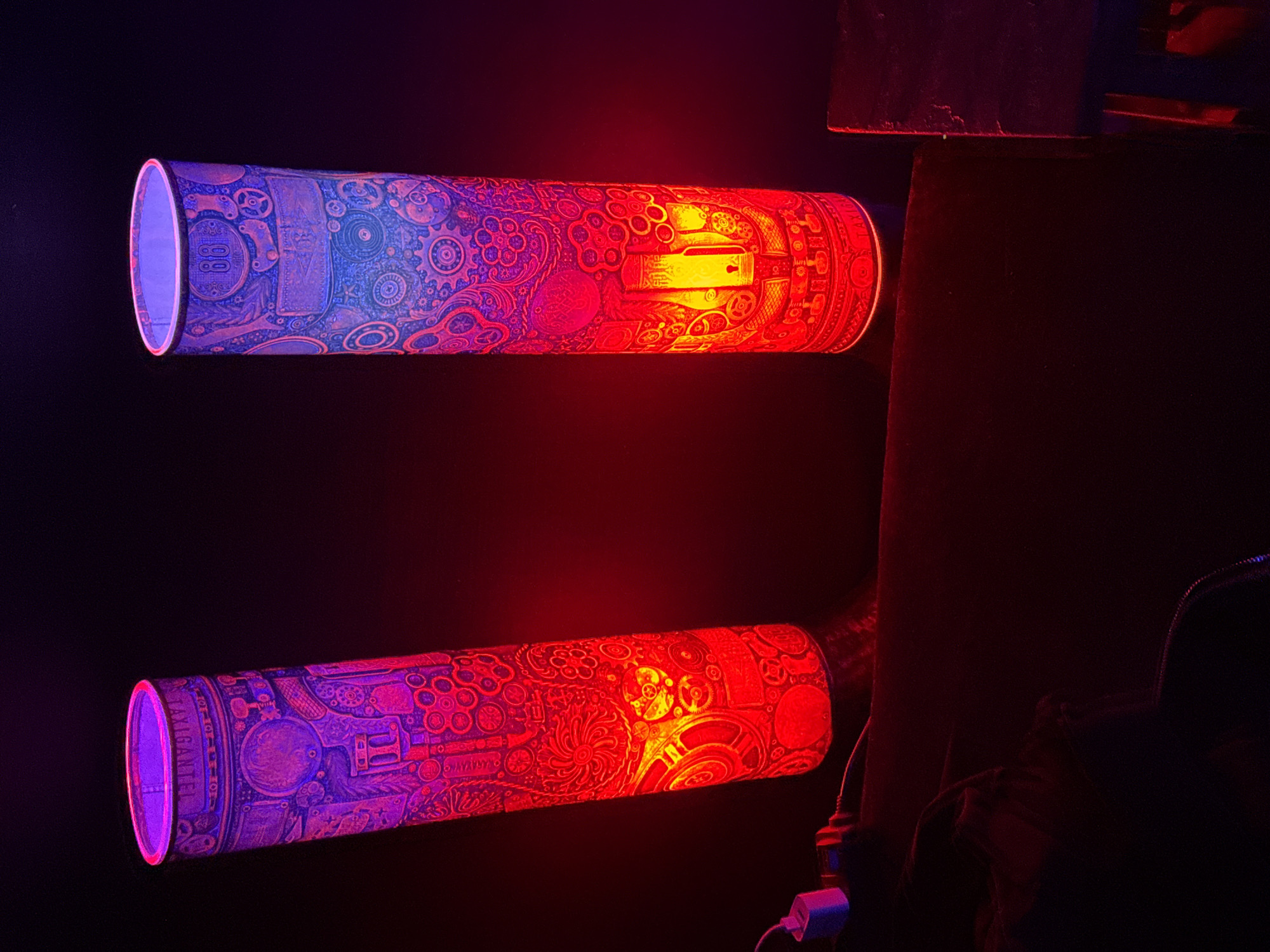 Two illuminated, cylindrical lamps with intricate, possibly steampunk-inspired designs are mounted on a dark wall. The light emanating from the left side of the lamps is primarily purple, transitioning to red on the right. The designs on the lamps appear to include gears, dials, and other mechanical components. The lamps are positioned horizontally, one above the other. Wires and a power adapter are visible near the lower lamp. The location is unknown.