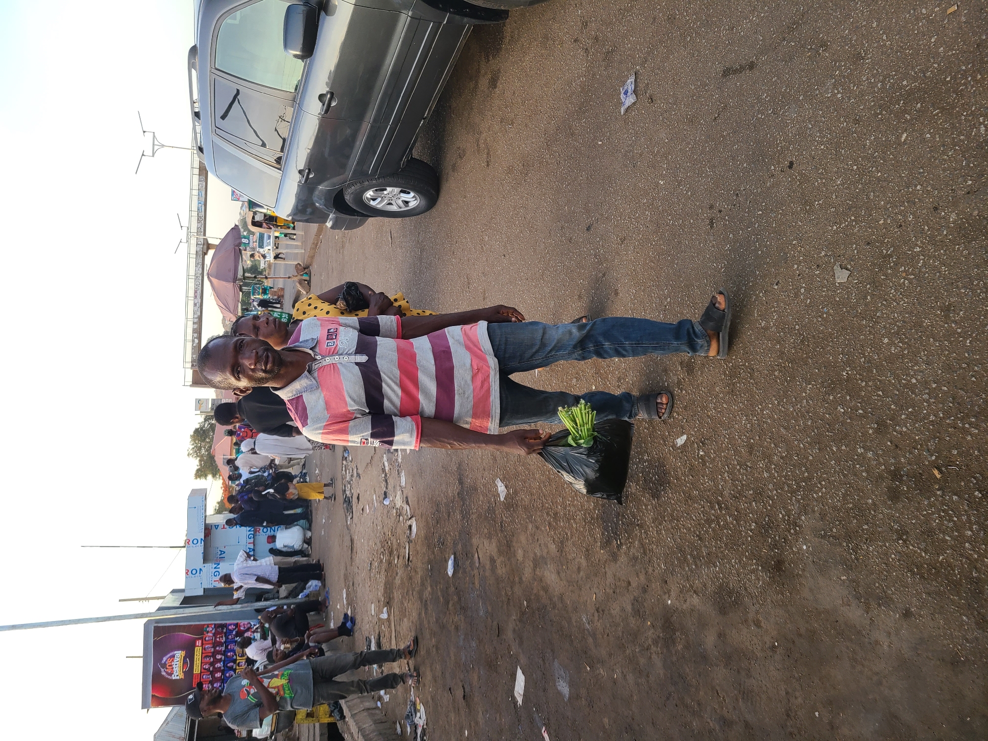 This is a vivid street scene capturing daily life in what appears to be a bustling market or roadside area in Jos, Nigeria. The image is taken at eye-level, presenting a dynamic view of people and activities.
In the foreground, a man stands prominently, looking directly at the camera with a neutral, slightly smiling expression. He is dressed in a striped short-sleeve polo shirt with alternating bands of pink, white, and a darker shade (possibly gray or maroon), blue jeans, and dark open-toed sandals. In his left hand, he holds a black plastic bag filled with fresh green vegetables, possibly greens or okra, indicating he has either just made a purchase or is selling. Behind him, partially obscured, is another person, likely a woman, wearing a yellow top with black polka dots.
The setting appears to be a busy street. A dark grey SUV is parked on the left side of the frame. The ground is a mix of asphalt and dirt, showing signs of wear and scattered litter, including pieces of paper and plastic. In the midground and background, numerous other people are visible, some walking, others gathered around market stalls or engaging in conversations. A purple umbrella suggests a vendor's stall or a shaded waiting area.
To the left, a large billboard is visible, partially displaying "LAGOS FESTIVAL" and an image of a bottle, along with multiple smaller portrait photos. Other signs and structures with partial text like "IRON" and numbers like "82" can be faintly seen in the background. The sky above is clear and bright, suggesting a sunny day, likely late morning or early afternoon, given the warm light and lack of harsh shadows. The overall atmosphere is lively and typical of a busy urban environment or market in the region.