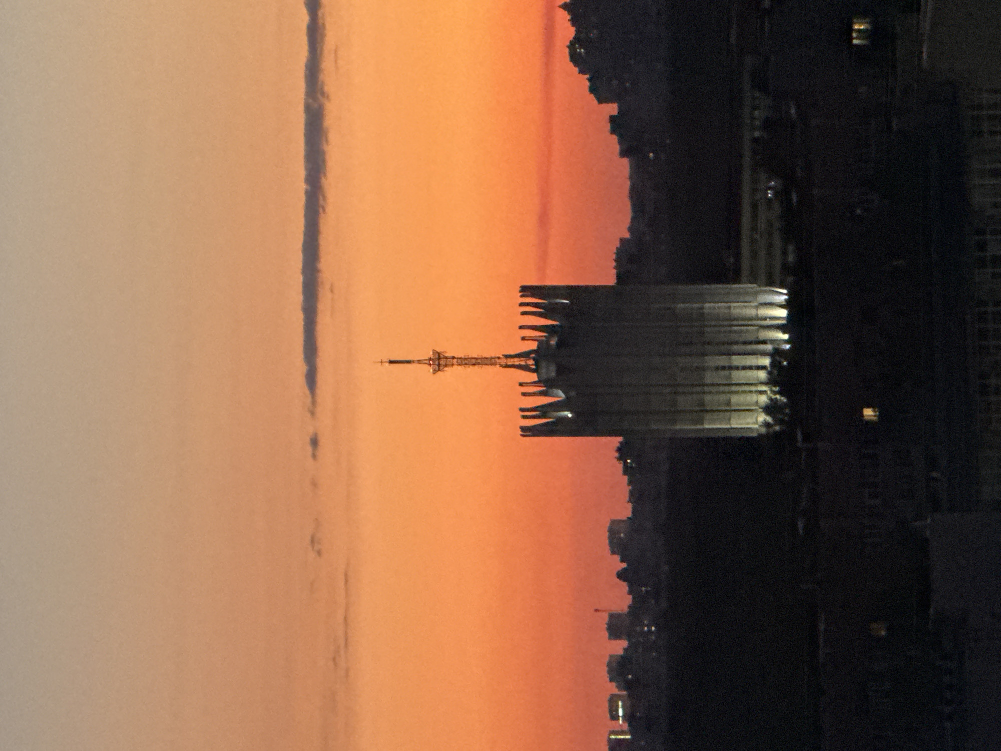 This image presents a twilight cityscape, likely at sunset, in Moscow, Russia. The central and dominant subject is the illuminated Main Building of Moscow State University. This tall, light-colored edifice features a distinctive multi-tiered upper section with prominent, vertical, fin-like architectural projections, which are brightly illuminated from below by artificial white lights. A slender spire, topped with an antenna-like mast and a radiant element, crowns the structure, also catching ambient light.
The sky above displays a vibrant gradient of orange and red, indicating a sunset, transitioning to a duller orange-grey at the top. Several elongated, dark cloud formations are visible, including a prominent horizontal one across the mid-upper section of the sky.
In the mid-ground, a dark, silhouetted urban skyline stretches across the horizon, showcasing various building profiles. A distinct red construction crane is visible on the left side of this silhouette. The foreground consists of dark, indistinct building and tree silhouettes, with a few discrete yellow lights visible in windows of structures in the lower-left and lower-right portions of the frame. No individuals are visible in the scene.