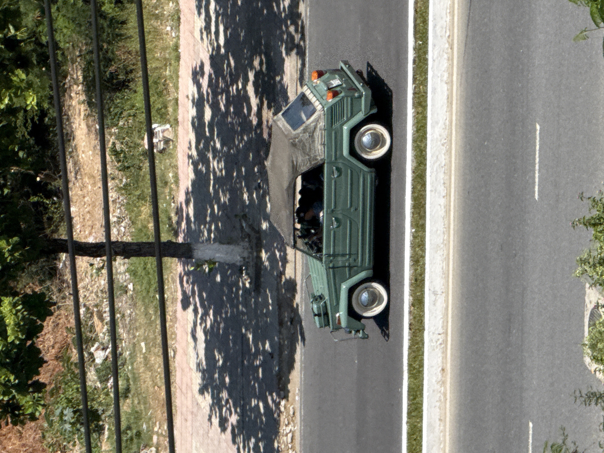 A high-angle shot captures a vintage, olive-green Volkswagen Thing convertible driving on an asphalt road in Nha Trang, Vietnam. The car, with its canvas top up, has white-rimmed wheels and chrome hubcaps. The interior appears occupied by at least one person. The road is bordered by a grassy strip with a white curb, and beyond that, a raised embankment with a metal railing. The background shows dense green foliage and a tree with its base painted white. The overall scene suggests a sunny day, with the shadows of trees cast across the road and embankment.
