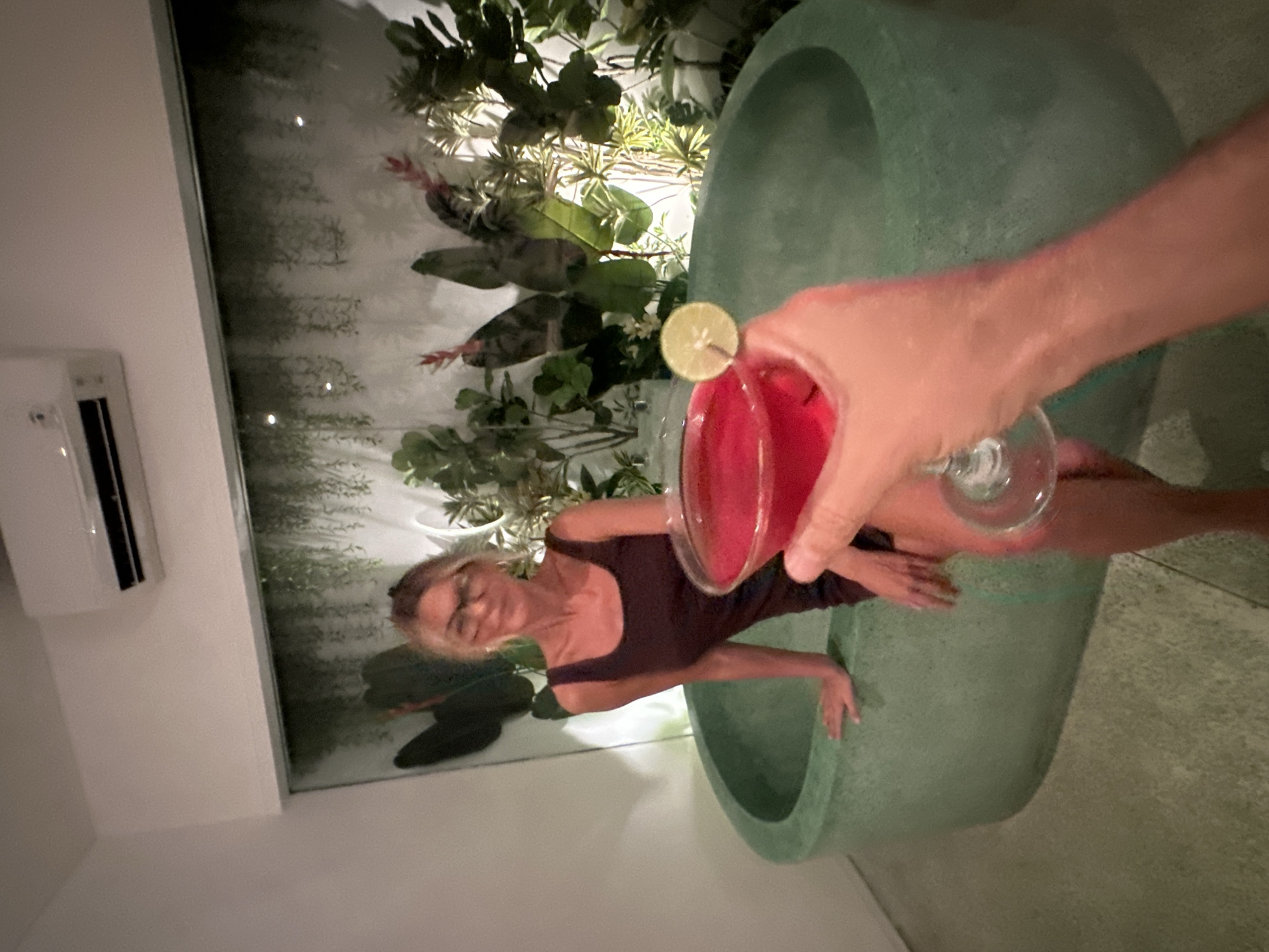 A photograph taken in North Kuta, Indonesia, presents a woman wearing a brown one-piece in a green tub. An arm holding a glass with a pink cocktail with a lime garnish is visible, held above her. The background includes a wall with plants. An air conditioning unit is mounted high on the left-hand wall.