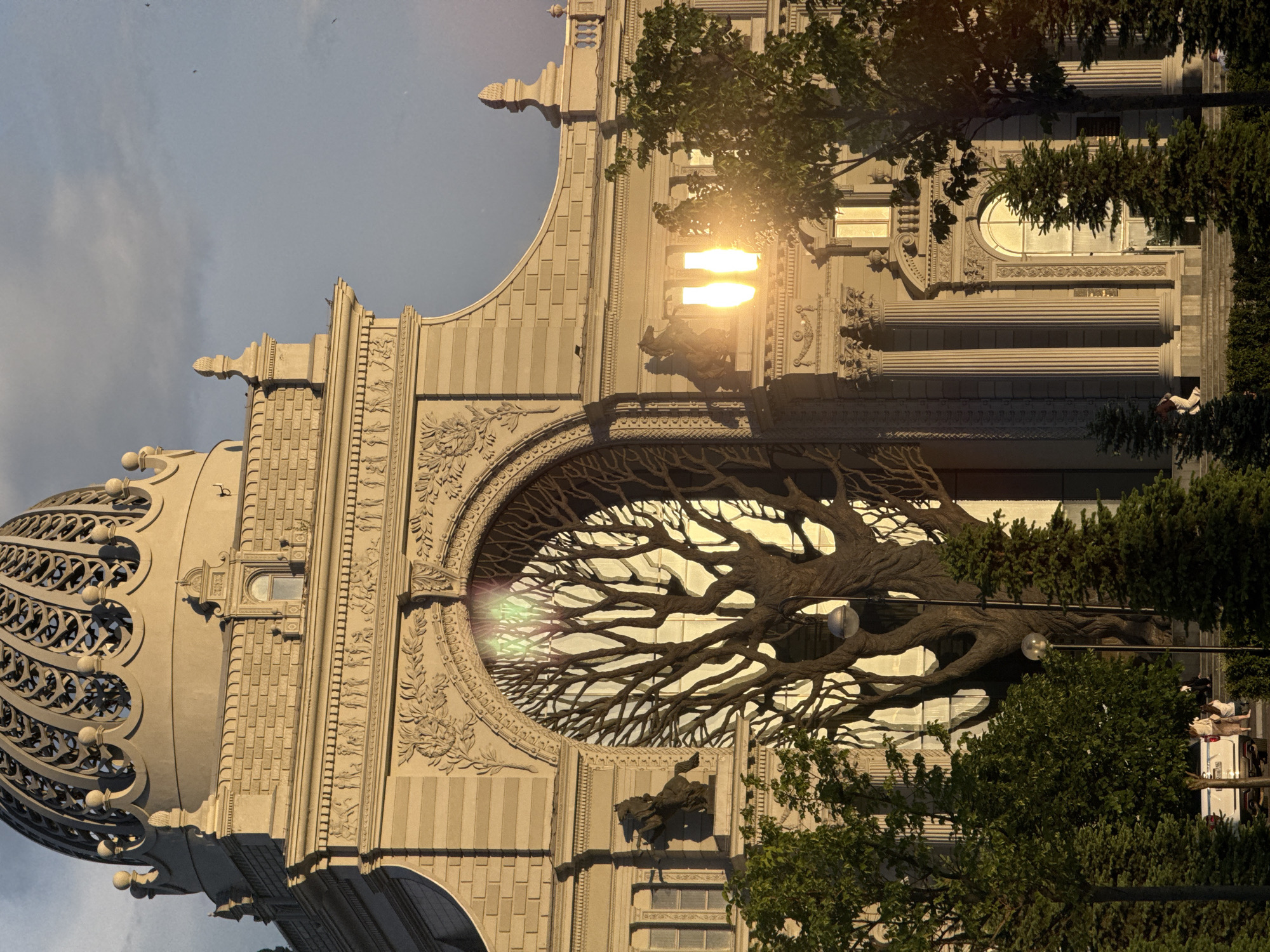 The image depicts a building with a distinctive architectural feature: a large, oval-shaped opening filled with a stylized tree sculpture made of brown material. The tree sculpture is positioned in front of a glass wall and features three glowing spheres within its branches. The building itself is constructed of light-colored stone and exhibits intricate detailing, including moldings, columns, and relief sculptures. The dome-shaped roof on the left side is adorned with an openwork pattern. Trees and foliage obscure portions of the lower section of the building. The lighting suggests it's either sunrise or sunset, and it is likely the Kazan Family Center in the city of Kazan, Russia.