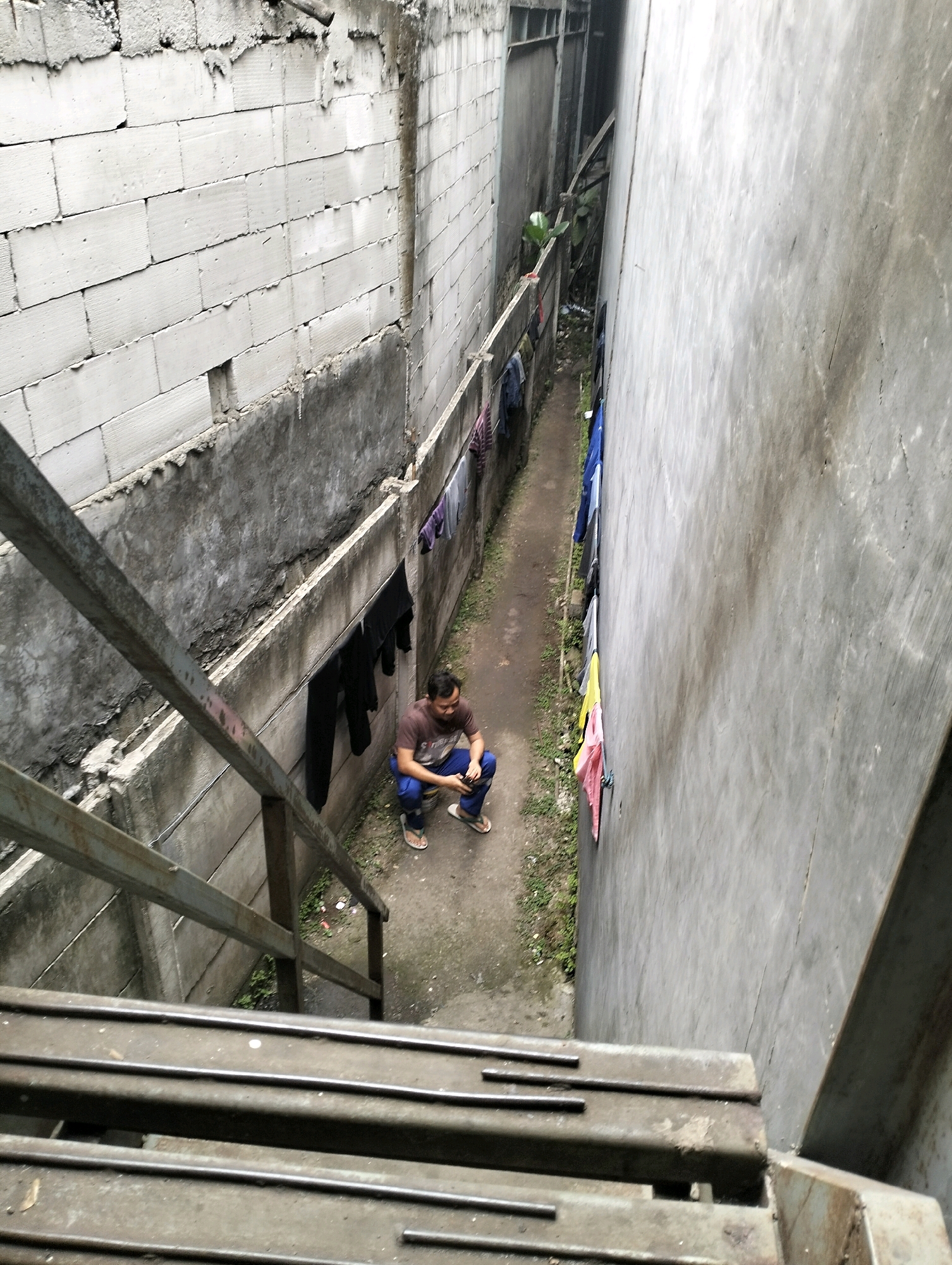 An overhead, eye-level shot shows a man squatting down in a narrow alleyway between two buildings in Gunung Sindur, Indonesia. The man, wearing a brown t-shirt and blue pants, is looking at his hands. The alleyway is covered in dirt and grass, with clothes hanging on a line above. The buildings on either side are made of concrete and brick. The building on the left has a brick wall and a concrete ledge, while the building on the right has a smooth concrete wall. In the foreground, a metal staircase leads down into the alleyway. The scene appears to be a residential area or a back alley.