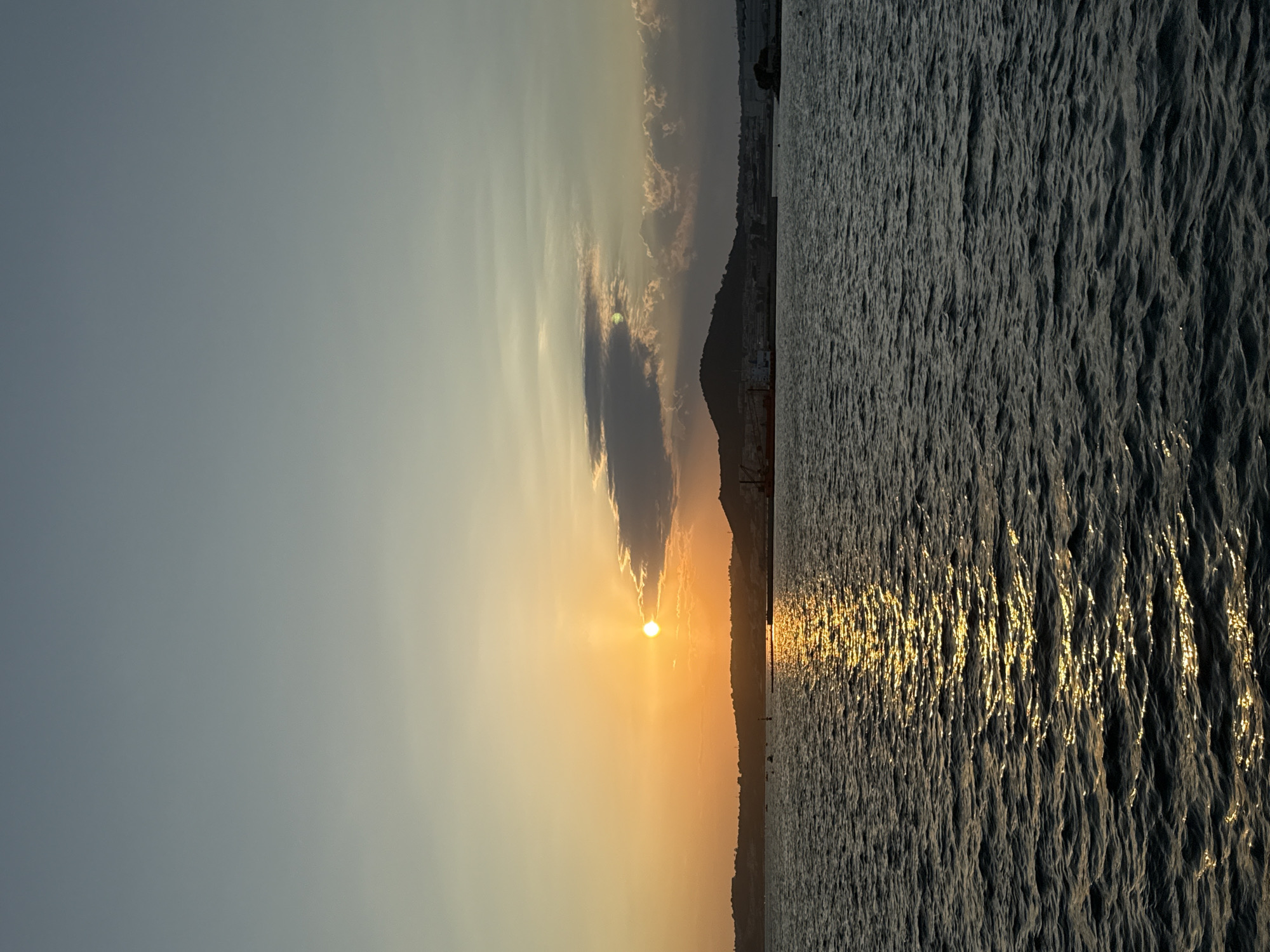 A sunset over the sea in Pozzuoli, Italy. The sun is low in the sky, casting a golden glow on the water. The sky is a mix of orange, yellow, and blue. In the distance, a silhouette of the city can be seen.