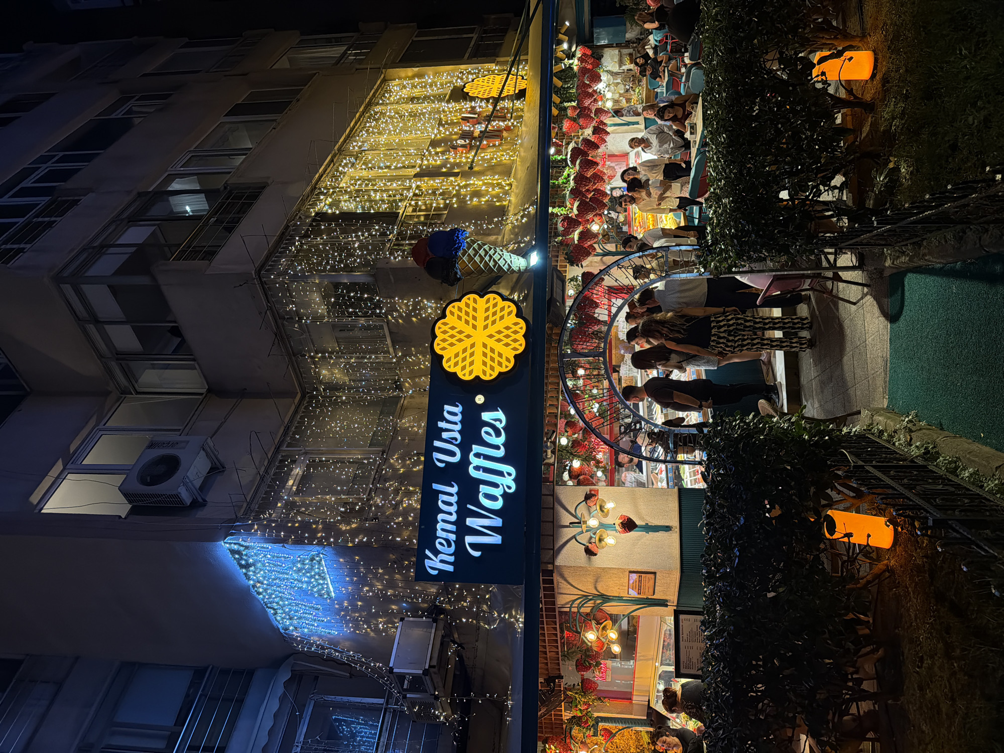 The image shows the exterior of "Kemal Usta Waffles" located in Kadıköy, Turkey, at night. The establishment is decorated with numerous string lights, some in blue and others in yellow, creating a festive atmosphere. A large sign with the name "Kemal Usta Waffles" is prominently displayed, featuring a waffle icon and an ice cream cone graphic. The front of the shop features a glass display case showcasing desserts and a counter where staff are serving customers. People are gathered both inside and outside the establishment, with some seated at tables. In the foreground, a low hedge with an orange lantern sits next to a black metal fence, and a green carpeted walkway leads towards the entrance. The building itself appears to be a multi-story structure with windows and balconies.