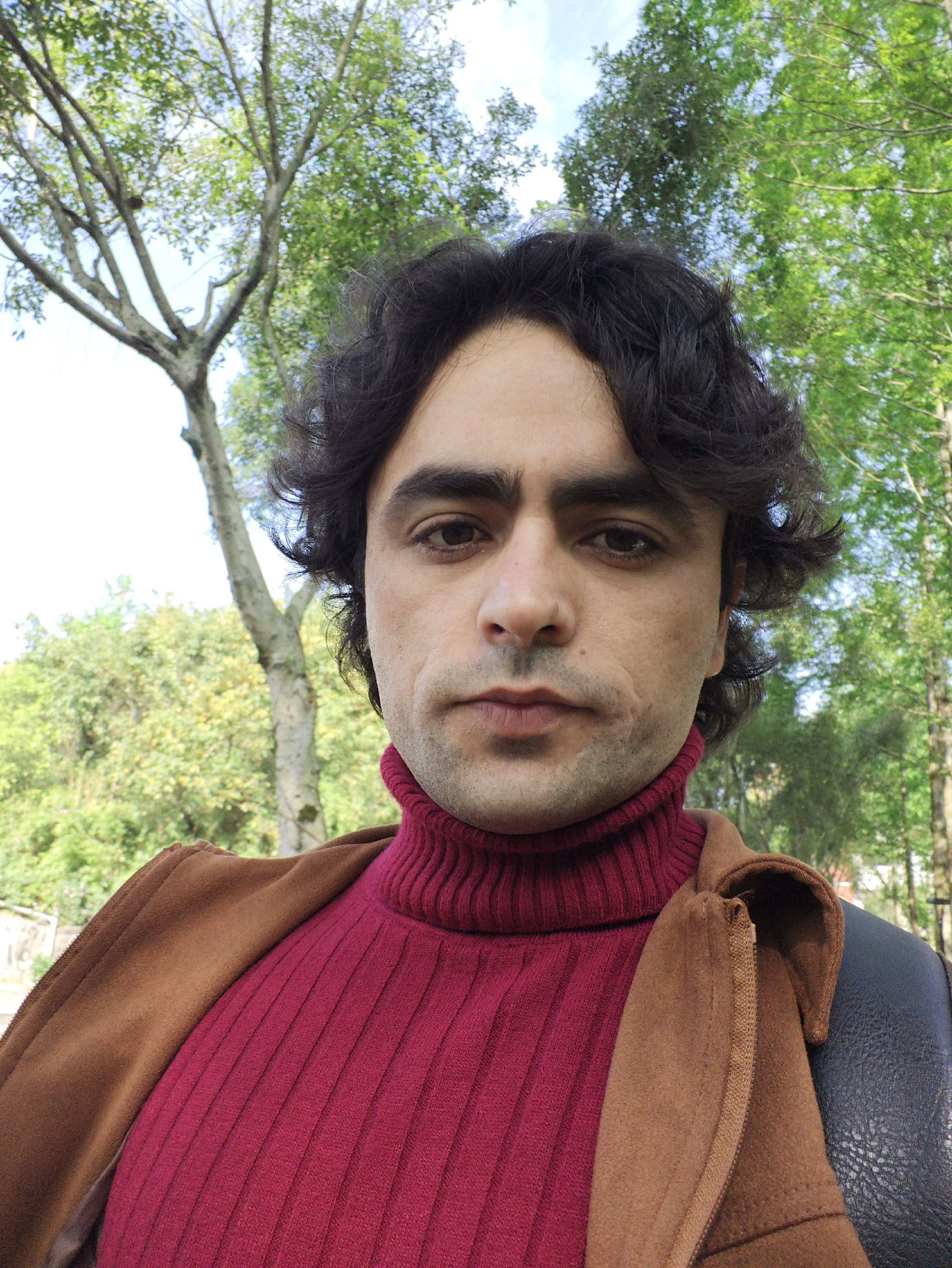 A medium shot of a man with dark hair and a maroon turtleneck sweater stands outdoors. He is wearing a brown jacket. The background features green trees. Based on prior context the location is Chongqing High-tech Zone, China.